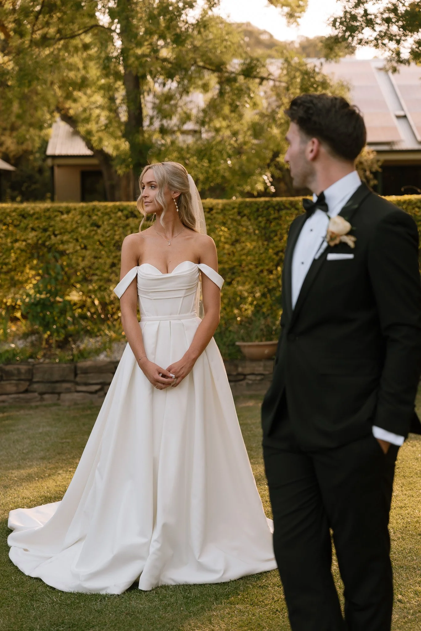 A bride and groom standing outdoors during sunset, with the bride wearing a white off-the-shoulder wedding gown and the groom in a black tuxedo with a bow tie, in a garden setting with trees and a house in the background.