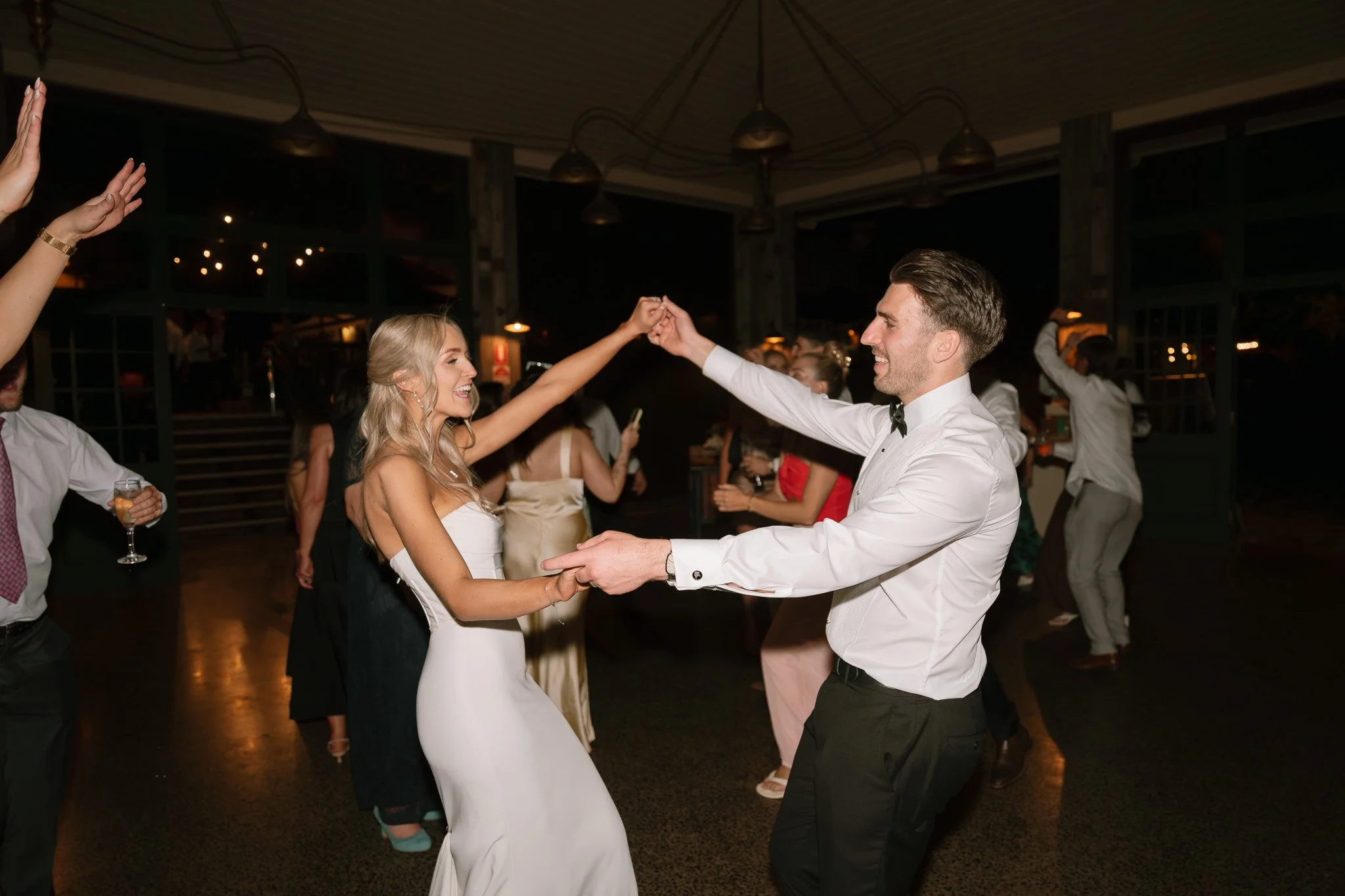A couple dancing at a wedding reception at night, holding hands and smiling. The woman wears a white dress, and the man wears a white shirt and black pants.