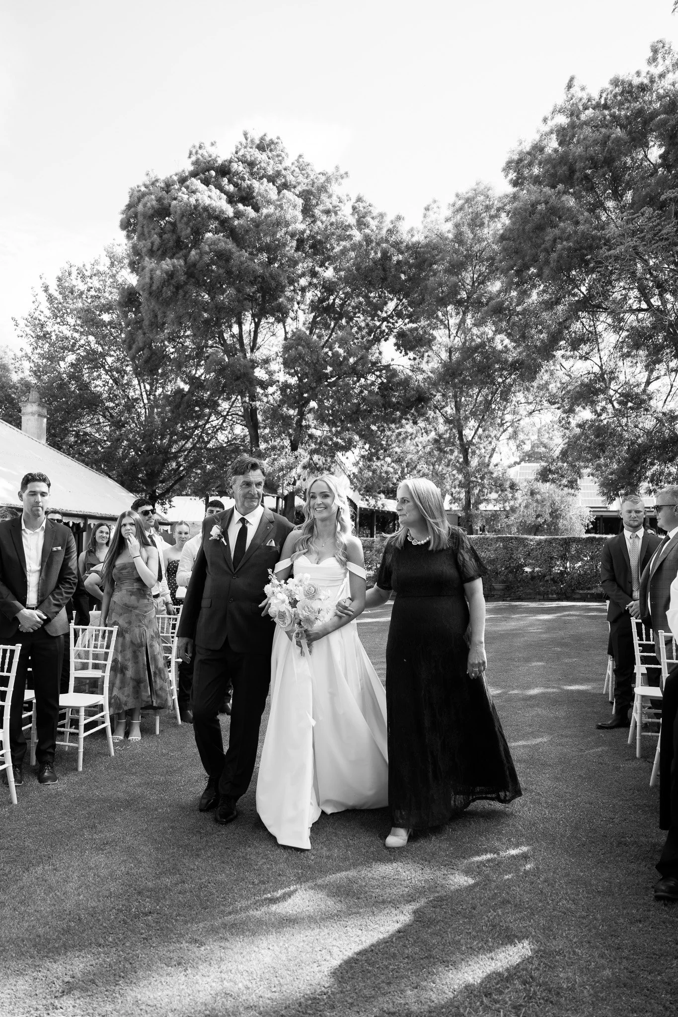A black and white photo of a bride walking down the aisle during a wedding ceremony, flanked by a man and a woman, outdoors with trees and guests in the background.