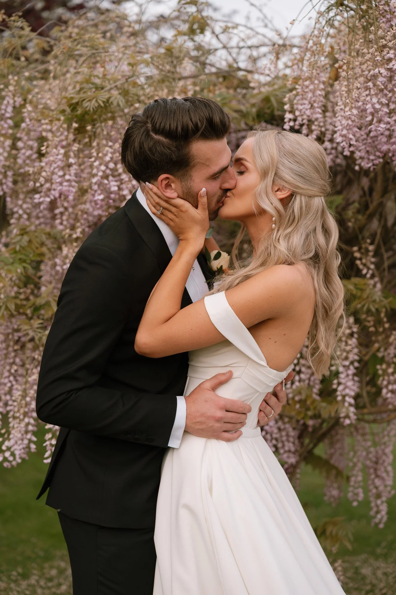 A bride and groom kissing outdoors surrounded by pink flowering trees, the groom wearing a black tuxedo and the bride in a white off-the-shoulder wedding dress.