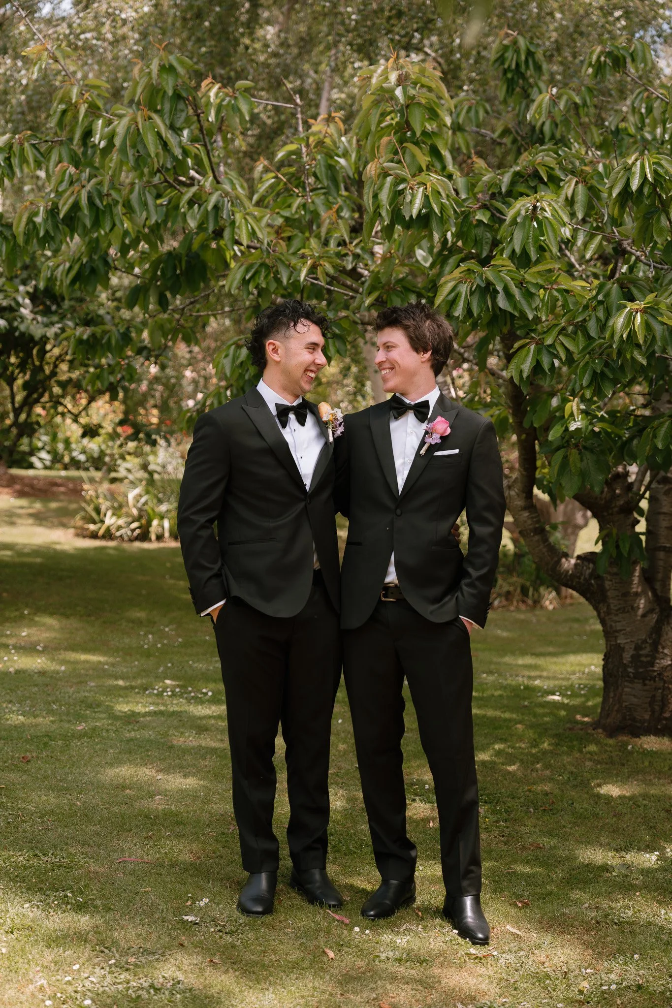 Two men in tuxedos standing arm in arm outdoors under a large green tree, smiling at each other, with a garden in the background.