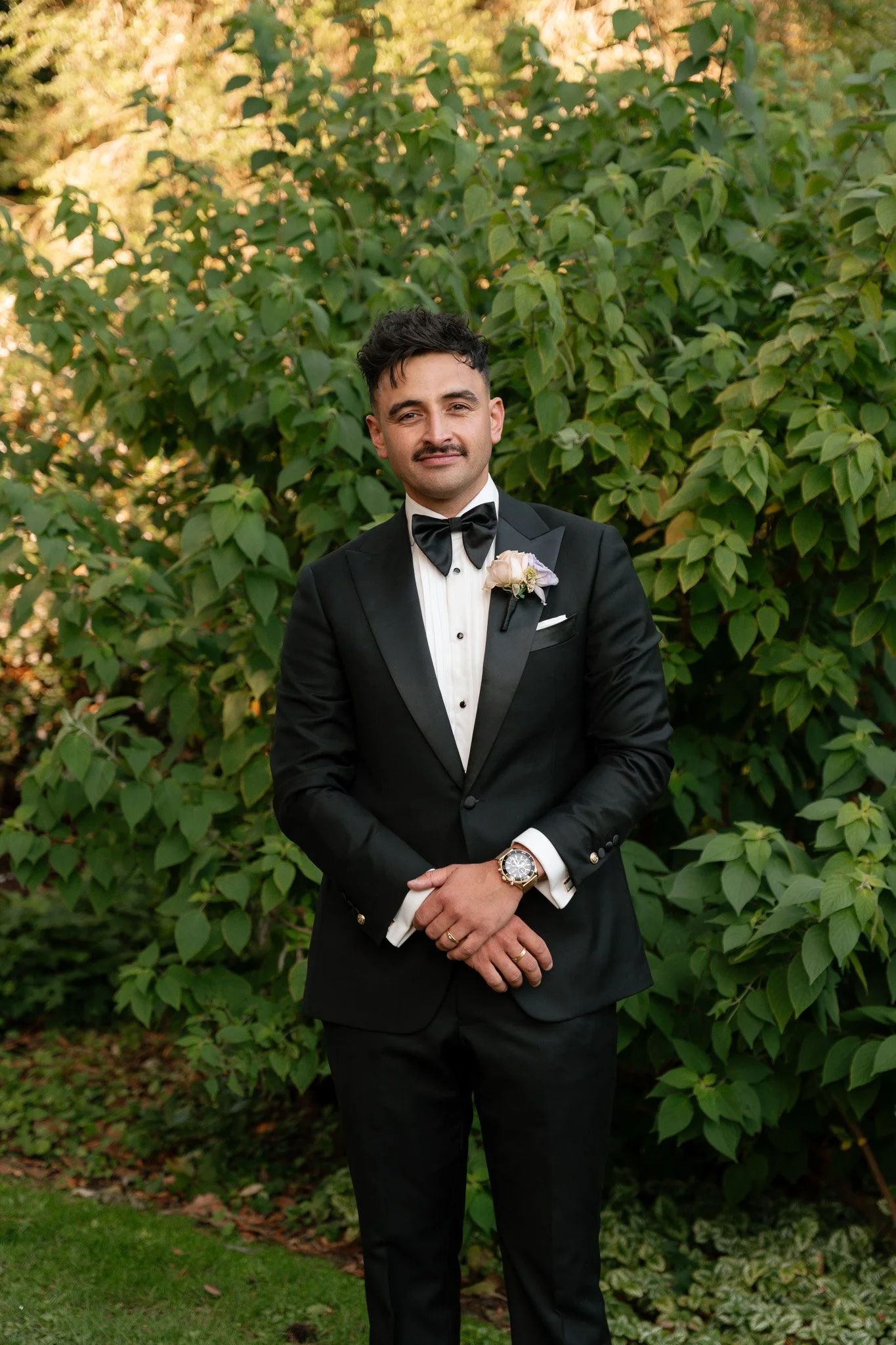 A man in a black tuxedo with a bow tie and boutonniere stands outdoors in front of green bushes, posing for a formal photograph.
