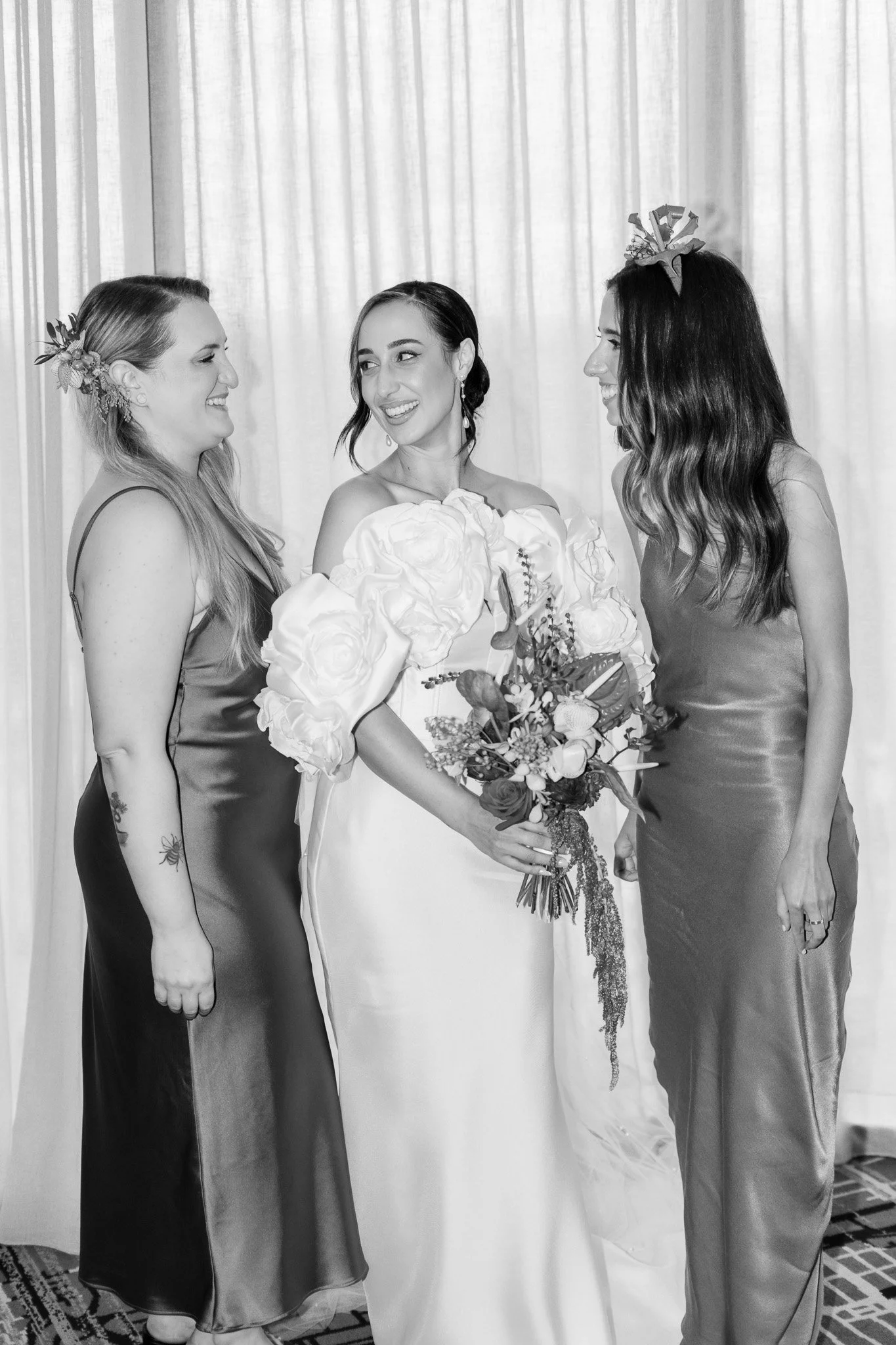 Three women, one in a wedding dress holding a bouquet, and two in bridesmaid dresses, standing and smiling at each other in front of curtains.