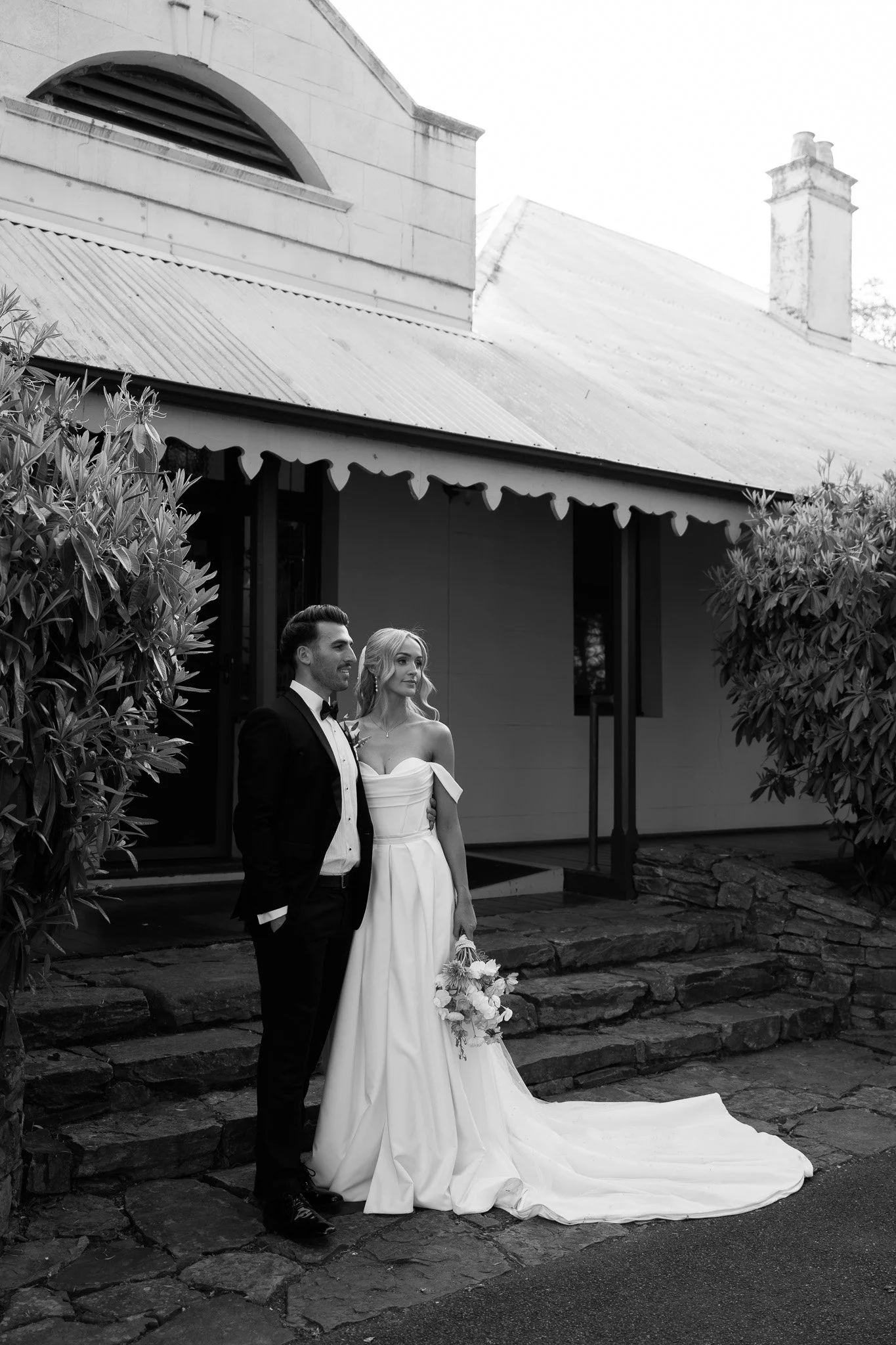 A black and white photograph of a bride and groom standing outside a house. The groom is in a tuxedo, and the bride is in a wedding gown holding a bouquet. They are posed on a stone pathway in front of a house with large windows and a corrugated meta