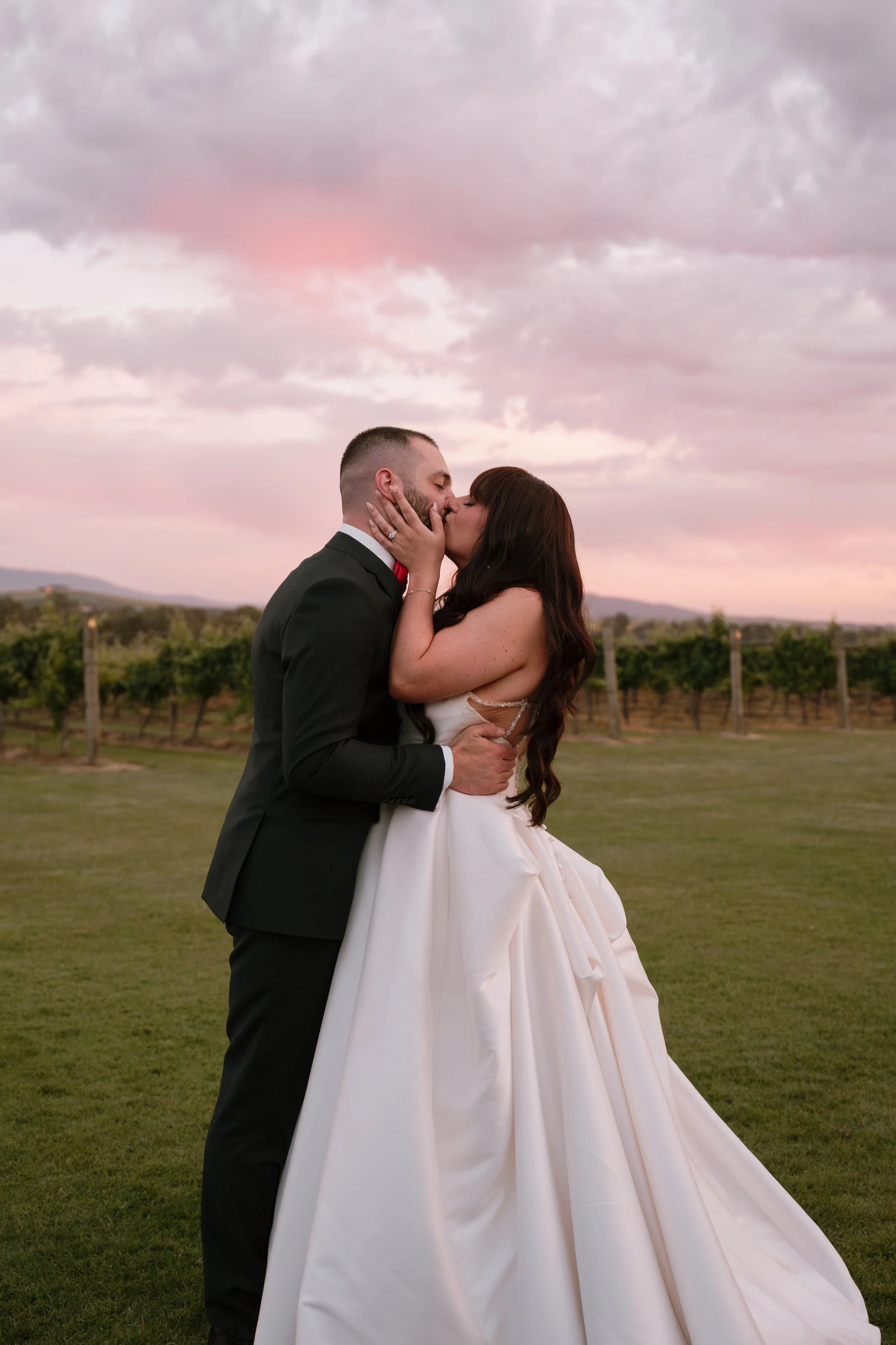 A newlywed couple sharing a kiss outdoors during sunset, with cloudy sky and vineyard in the background.