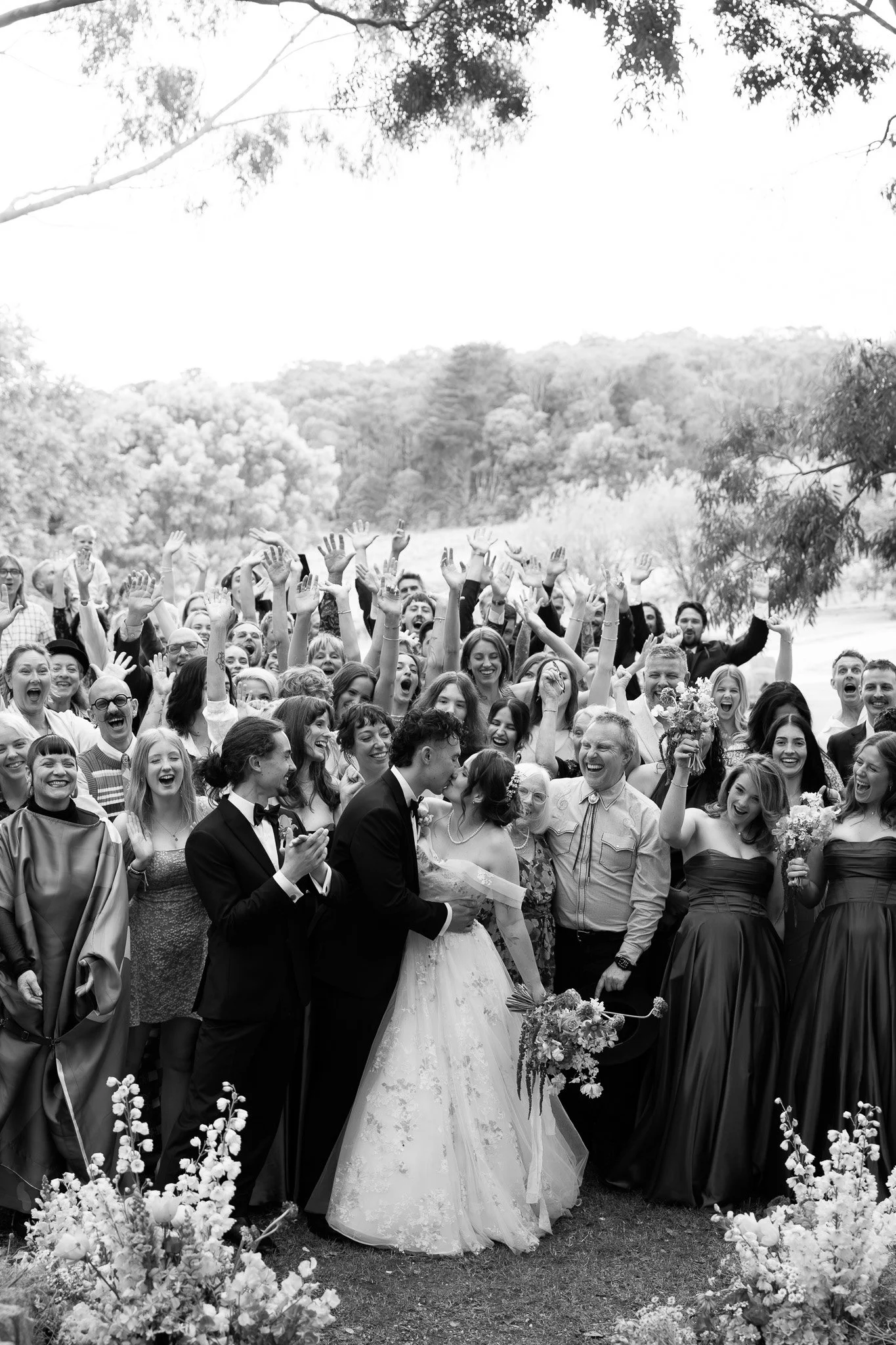 A black and white photo of a large outdoor wedding celebration with many guests cheerfully surrounding a bride and groom, who are kissing, with the bride holding a bouquet, people cheering and clapping, and trees in the background.
