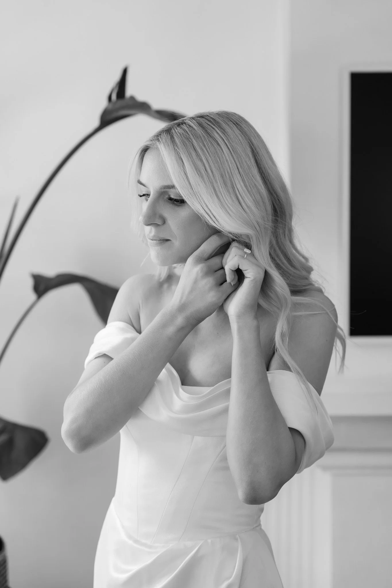 A woman with long blonde hair adjusting an earring, wearing a white off-the-shoulder dress indoors.