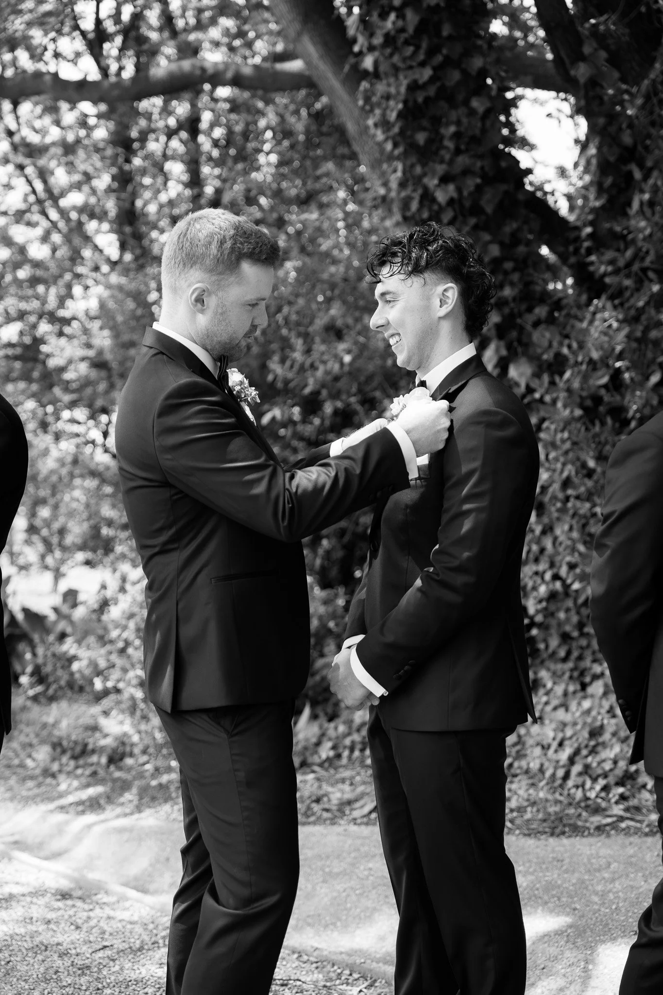 Two men in tuxedos, one pinning a boutonniere on the other's lapel, during a wedding ceremony outdoors, with trees and leaves in the background.
