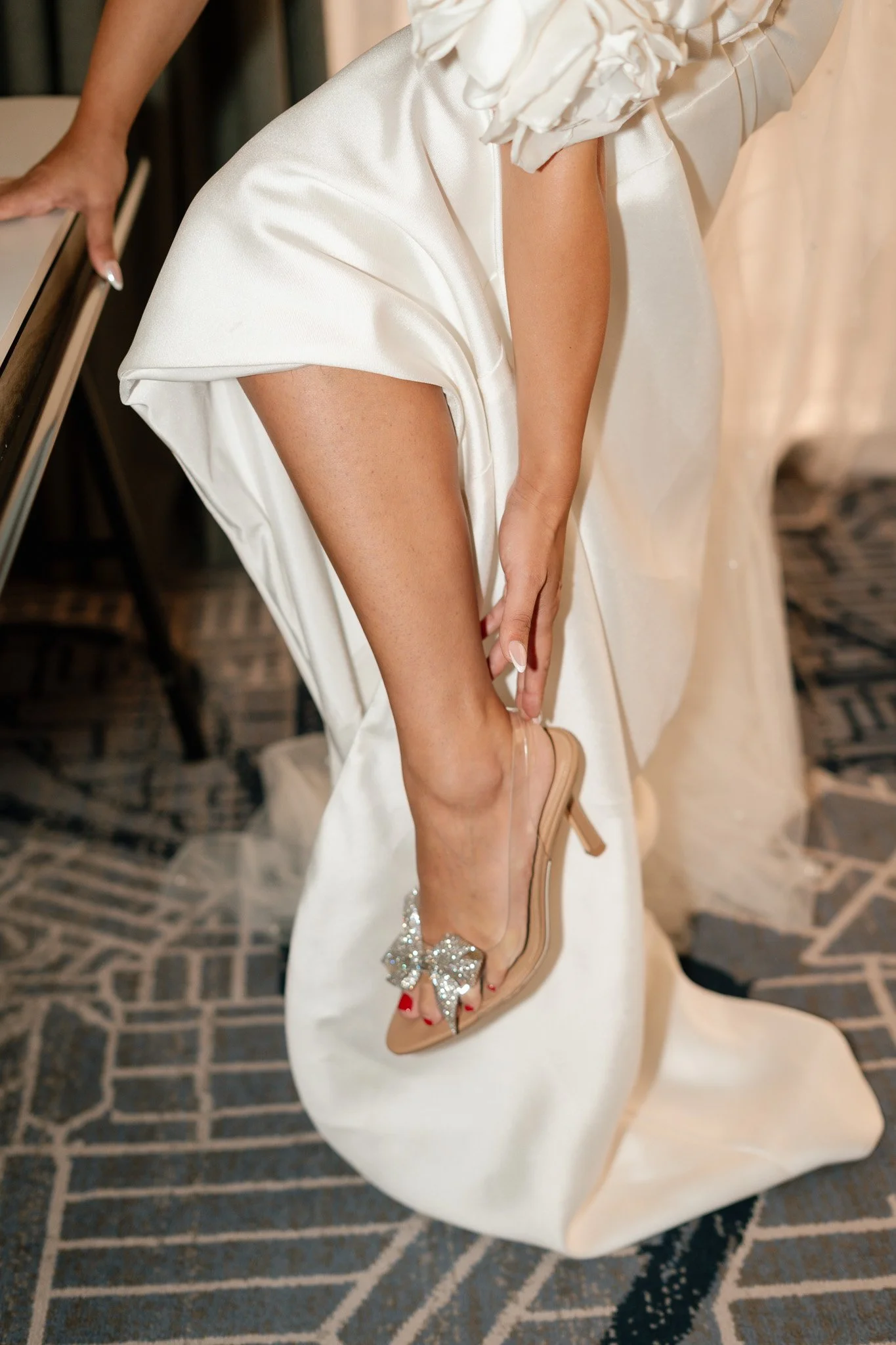 A woman in a white satin dress is adjusting her nude high-heeled sandal with a large rhinestone bow on the toe.