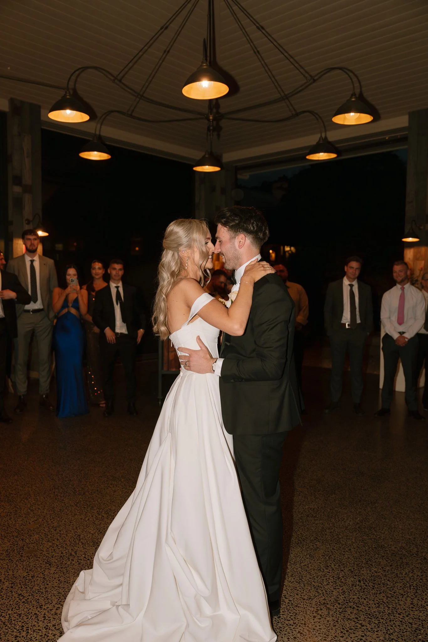 A bride and groom share their first dance at their wedding reception, surrounded by friends and family at night.