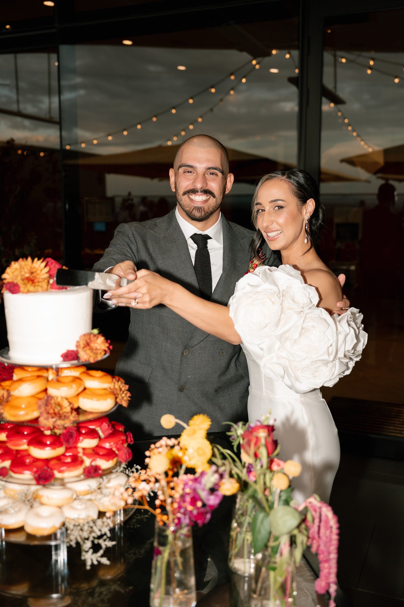 A couple cuts a wedding cake at their celebration, with a decorated table of flowers and desserts in front of them, and string lights in the background.