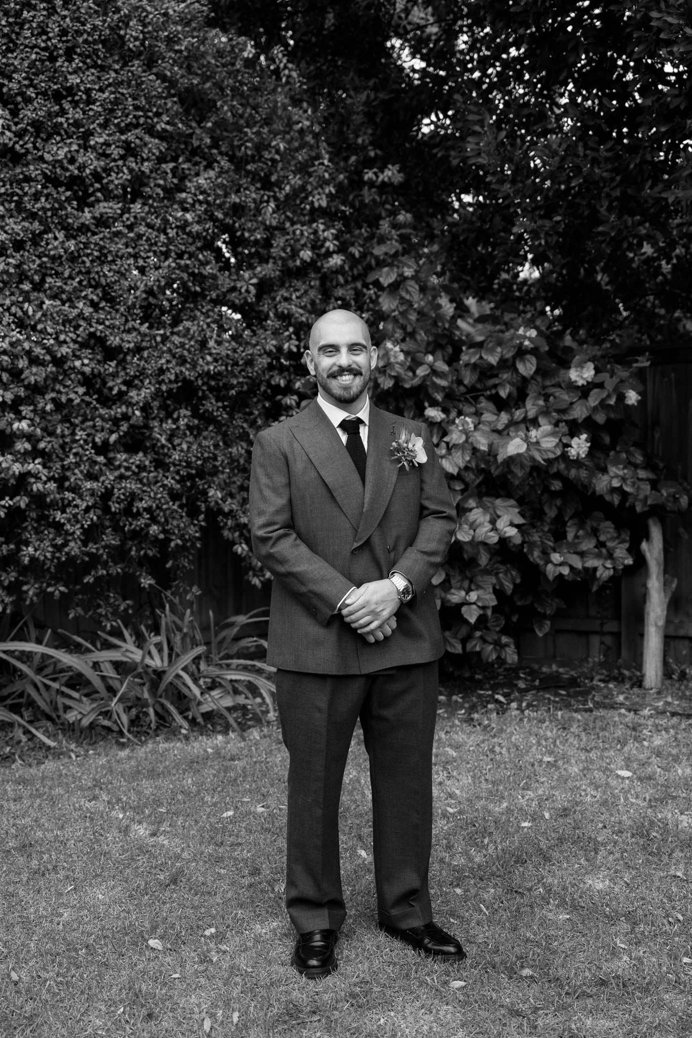 A man in a suit standing outdoors on grass with trees and bushes in the background, smiling at the camera.