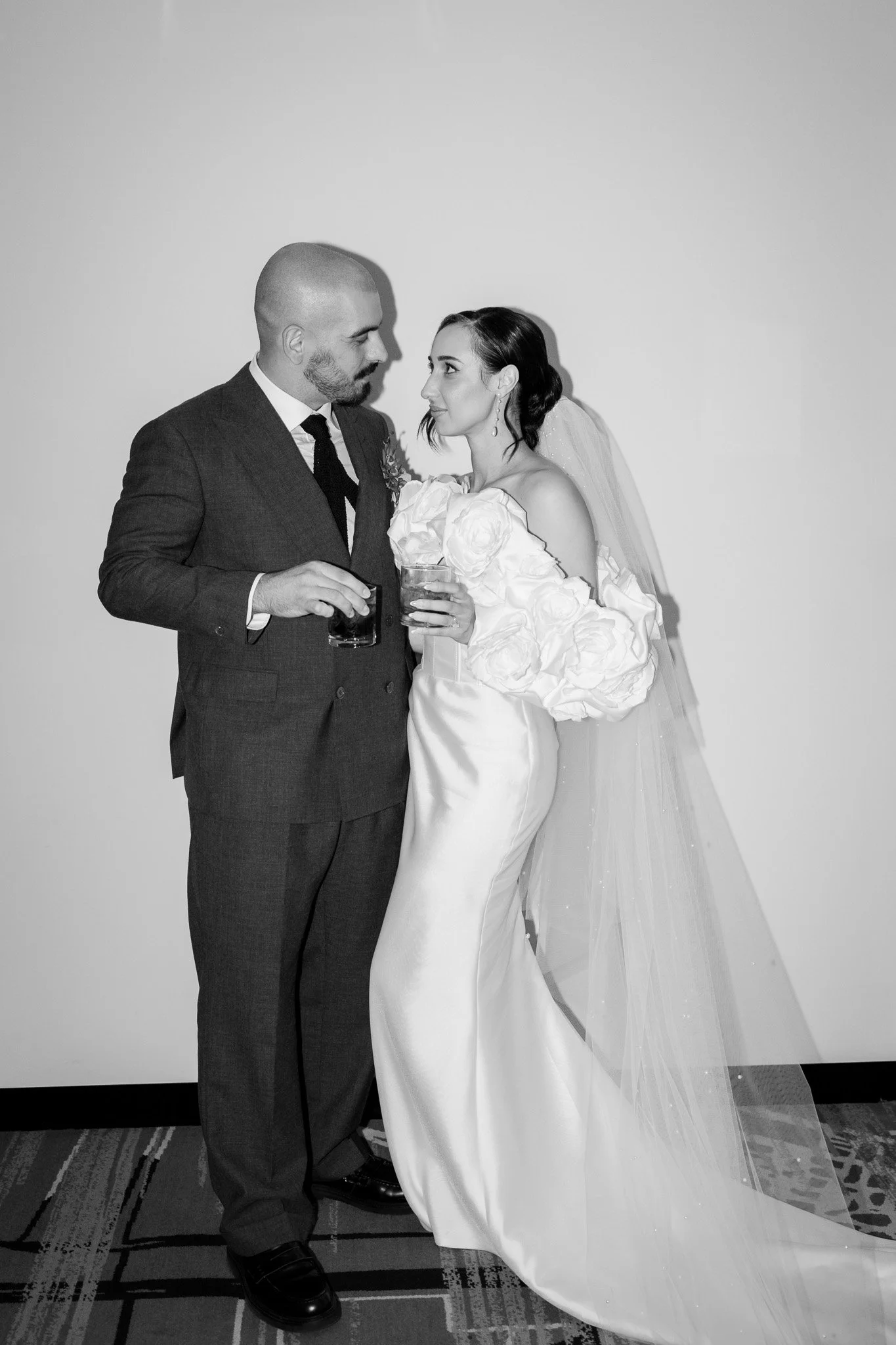 Black and white photo of a bride and groom standing close together, each holding a drink, looking at each other. The bride is wearing a satin wedding gown with floral embellishments and a veil, the groom is in a suit with a white shirt and tie. They 