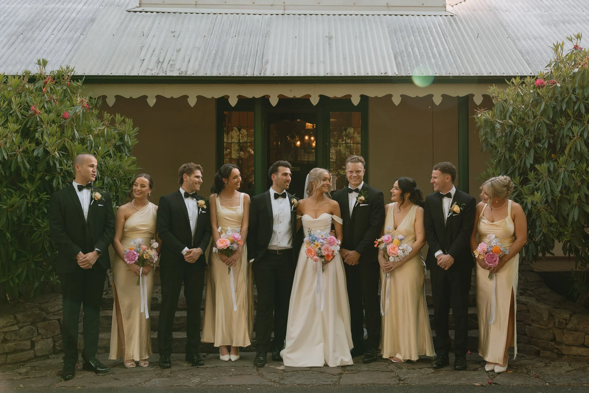 Wedding party with bridesmaids and groomsmen standing outside a building with a metal roof, smiling and holding floral bouquets.