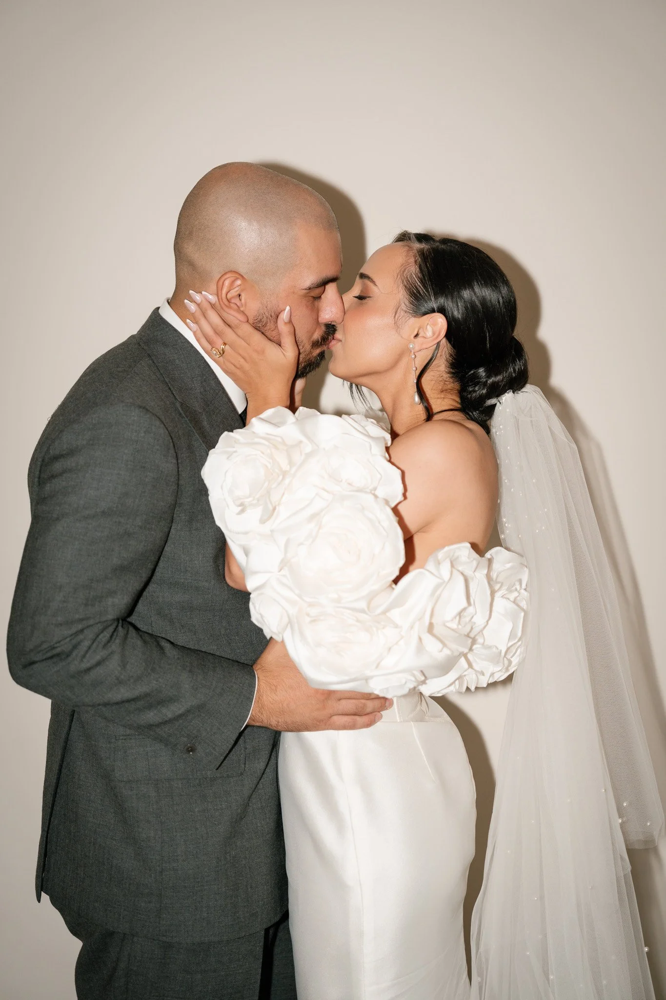 A bride and groom share a kiss during their wedding, with the bride dressed in a white gown with oversized puffed sleeves and a veil, and the groom in a gray suit, against a plain white wall.