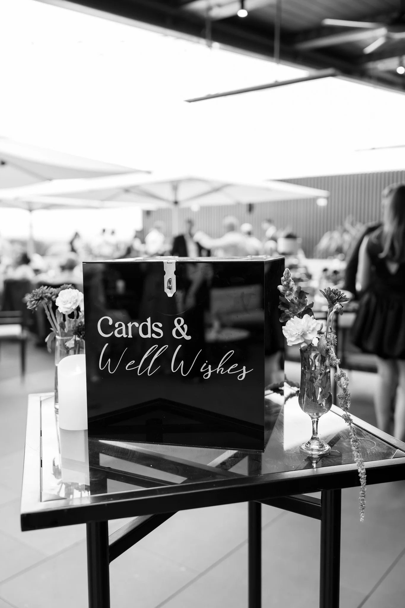 A black sign on a table reads 'Cards & Well Wishes'. The table also has a flower arrangement in a small vase and a white candle. People are in the background at an outdoor event with umbrellas and a roof overhead.