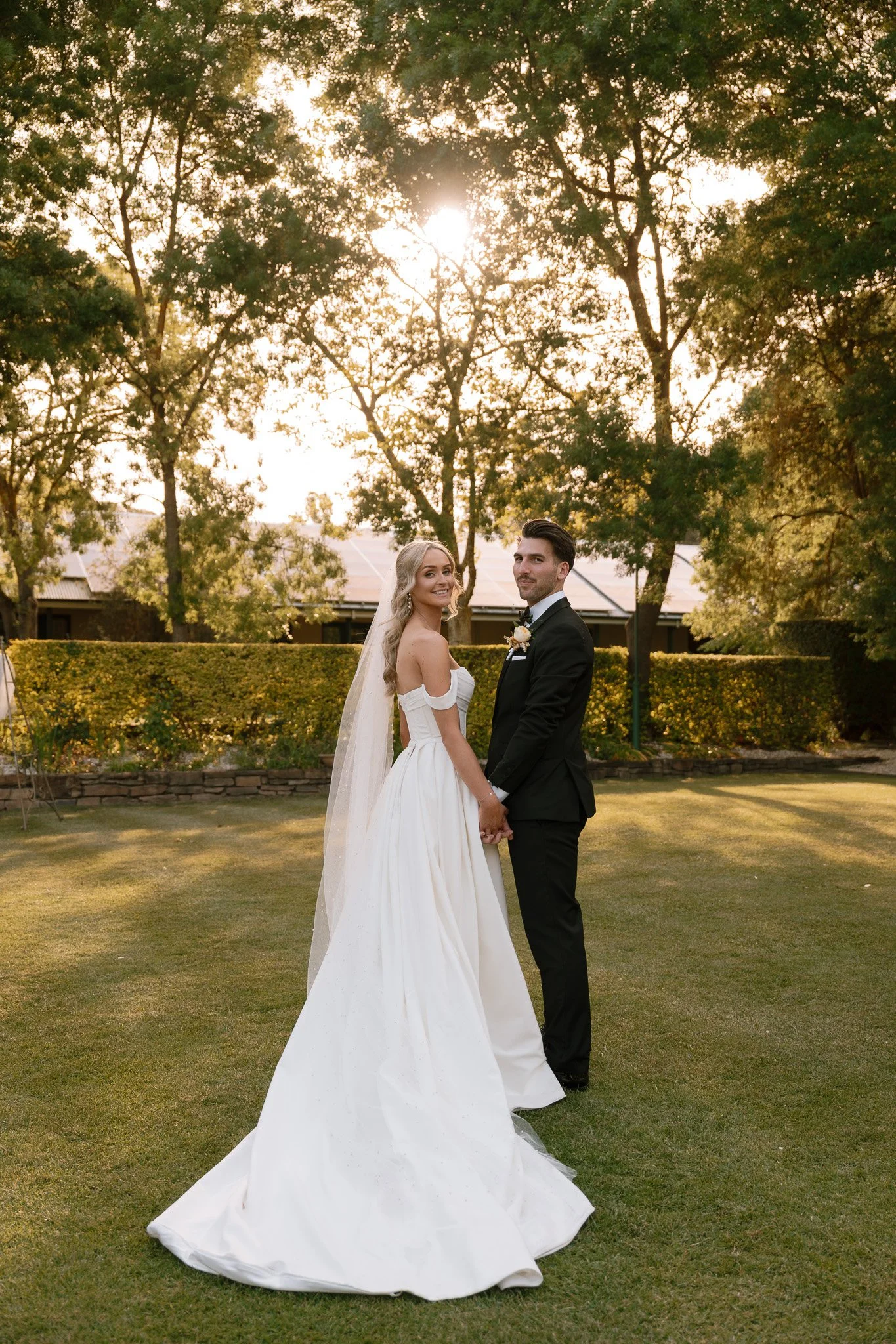 A newlywed couple holding hands and smiling at the camera outdoors during sunset, with a backdrop of trees and a hedge, in wedding attire.