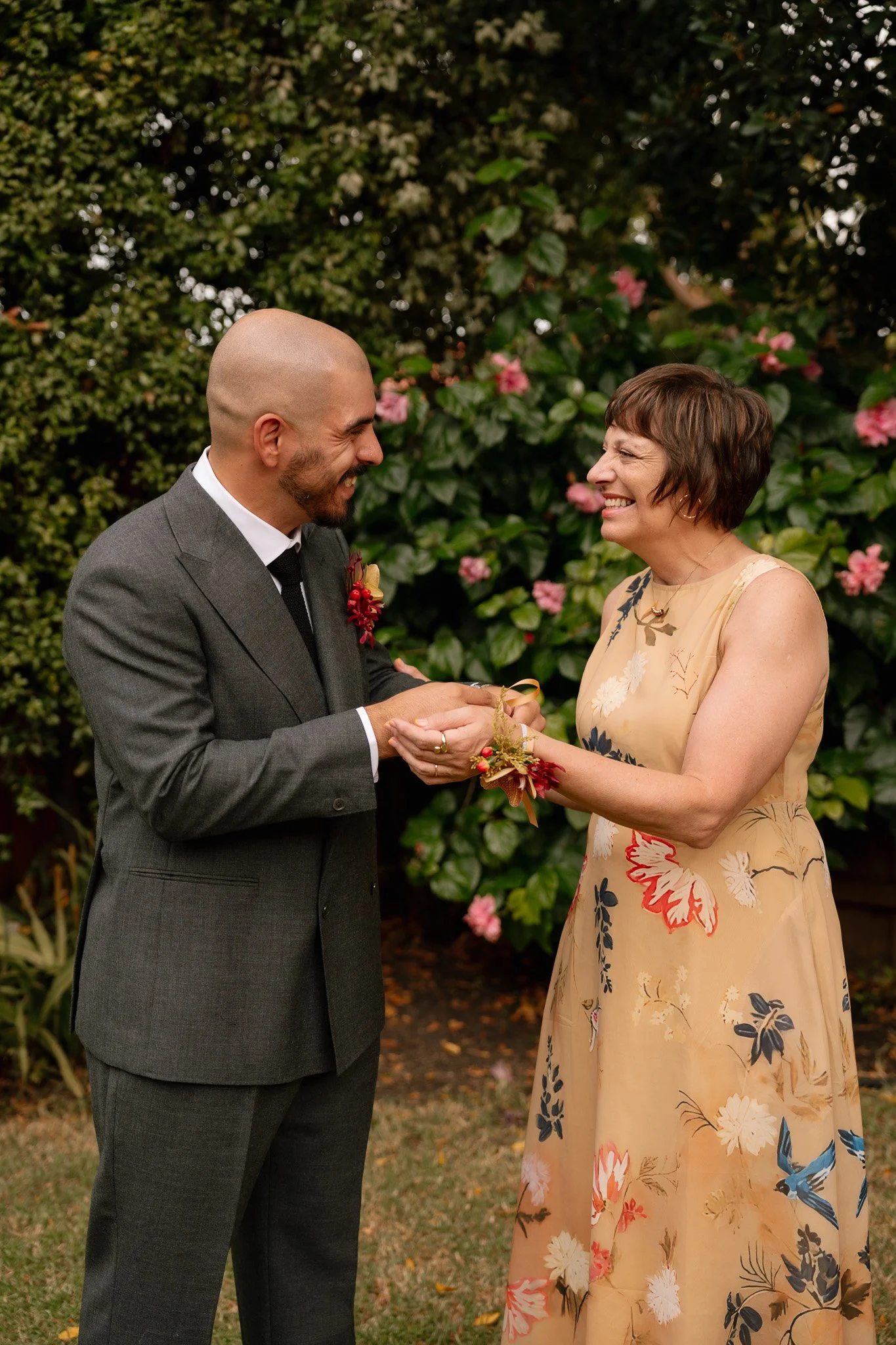 A man in a gray suit and a woman in a floral dress exchange rings outdoors, smiling, with pink flowers and green foliage in the background.