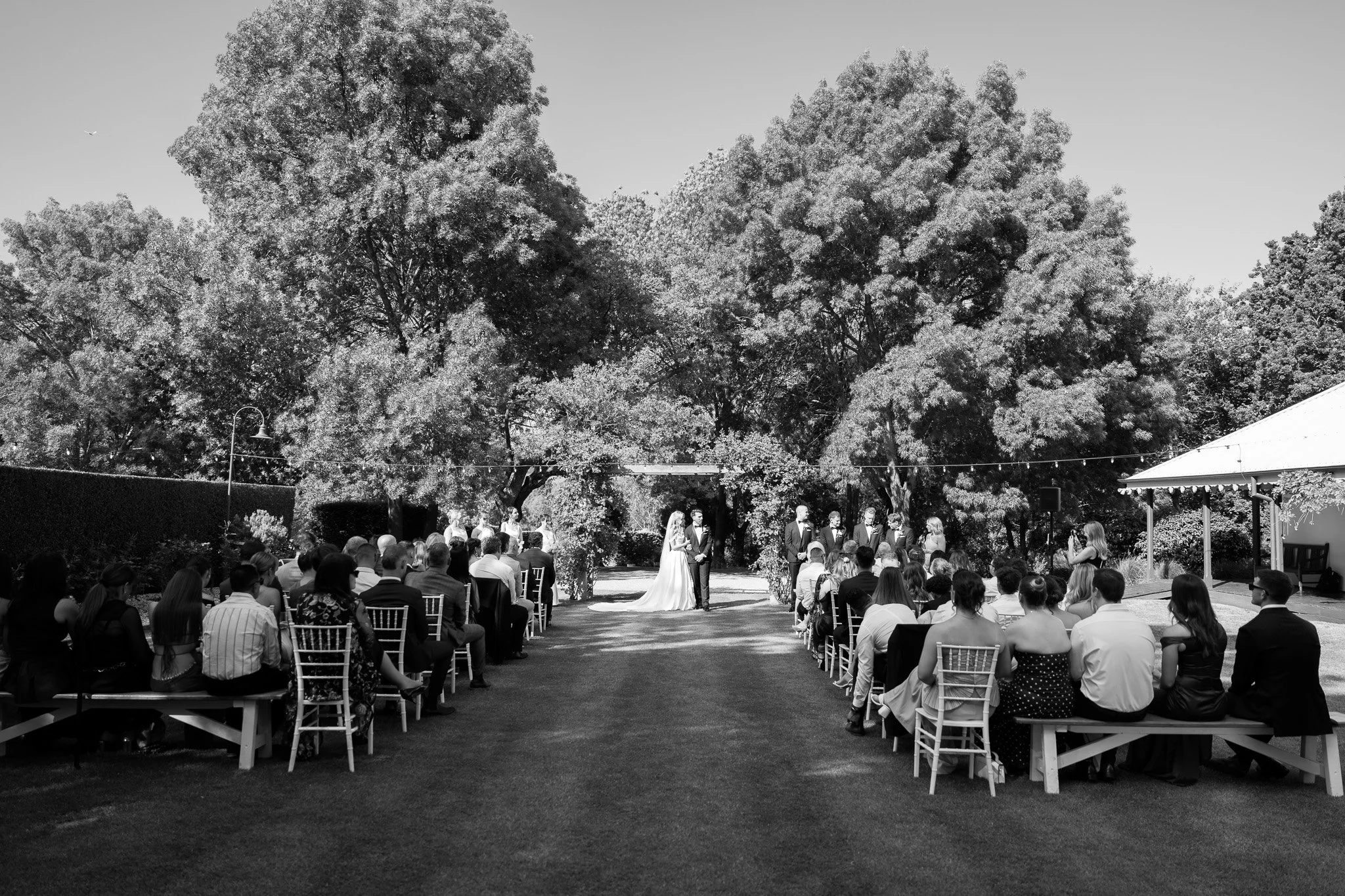 A black and white photograph of an outdoor wedding ceremony. The bride and groom stand in the center in front of an officiant, surrounded by trees. Guests are seated on either side in rows and benches on the grass, watching the ceremony.