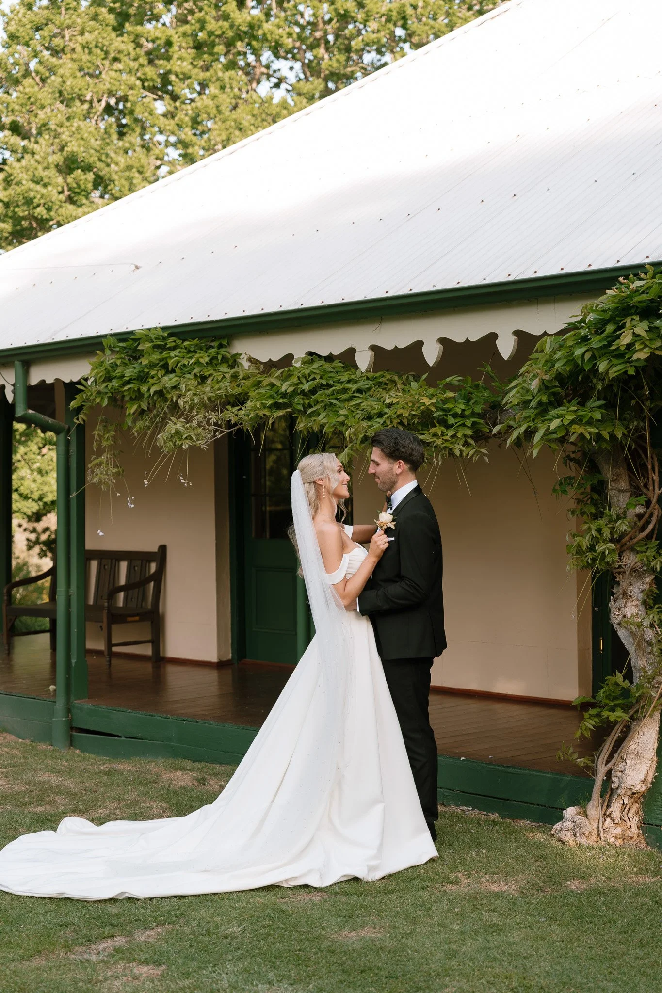 A bride and groom sharing a moment outdoors on their wedding day, standing on grass in front of a quaint building with green door and vine-covered porch.