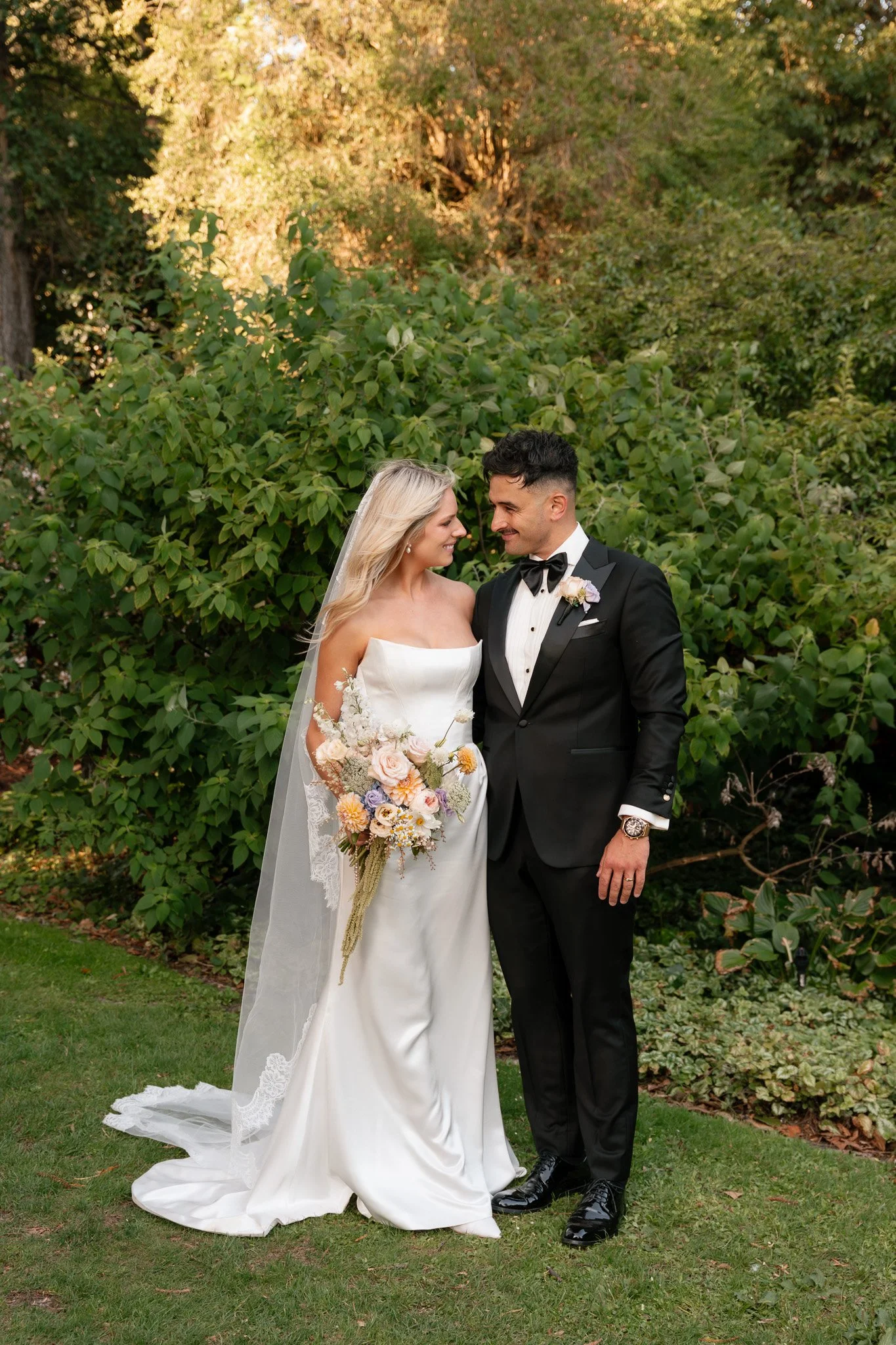 A bride and groom stand close together, smiling and looking into each other's eyes outdoors surrounded by green foliage, on their wedding day. The bride is holding a bouquet of pastel flowers and wearing a white strapless wedding gown with a veil. Th