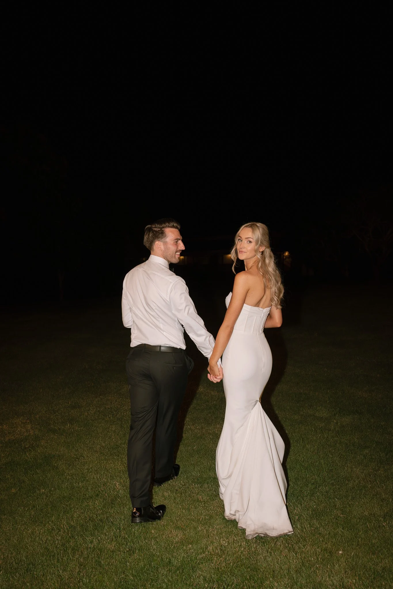A couple in formal attire holding hands outdoors at night, with a woman wearing a strapless white gown and a man in a white dress shirt and black pants, standing on grass.