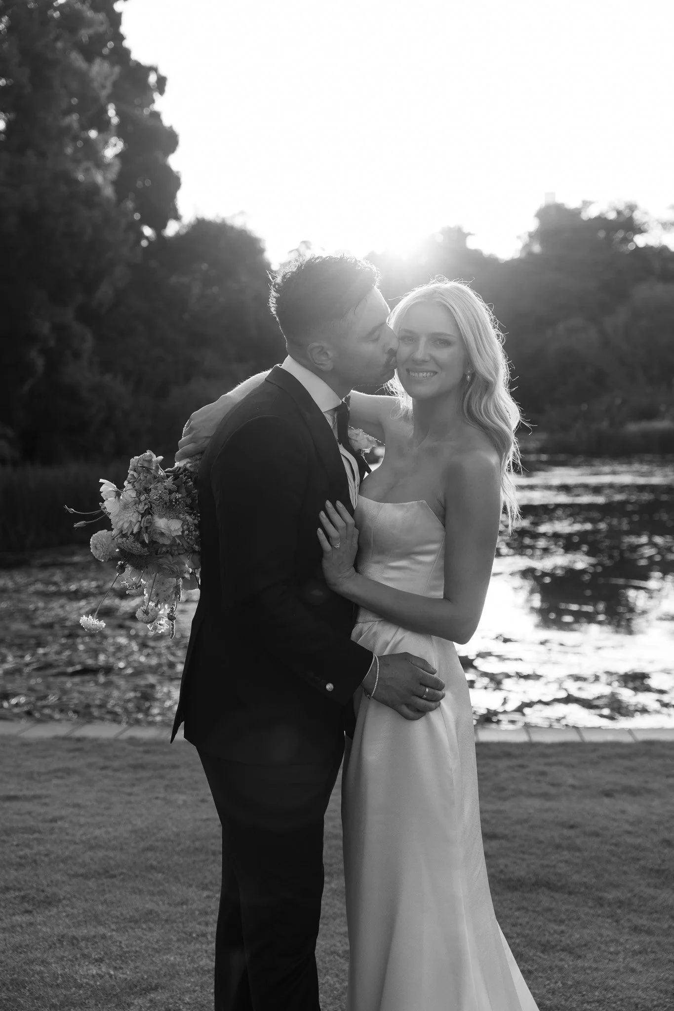 A black-and-white photo of a bride and groom at sunset near a lake, with the groom kissing the bride on the cheek and she smiling, holding a bouquet of flowers.