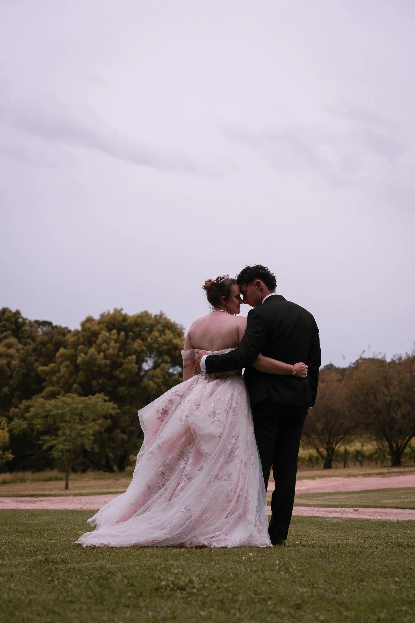 A couple in wedding attire embracing outdoors on a cloudy day, with trees in the background.