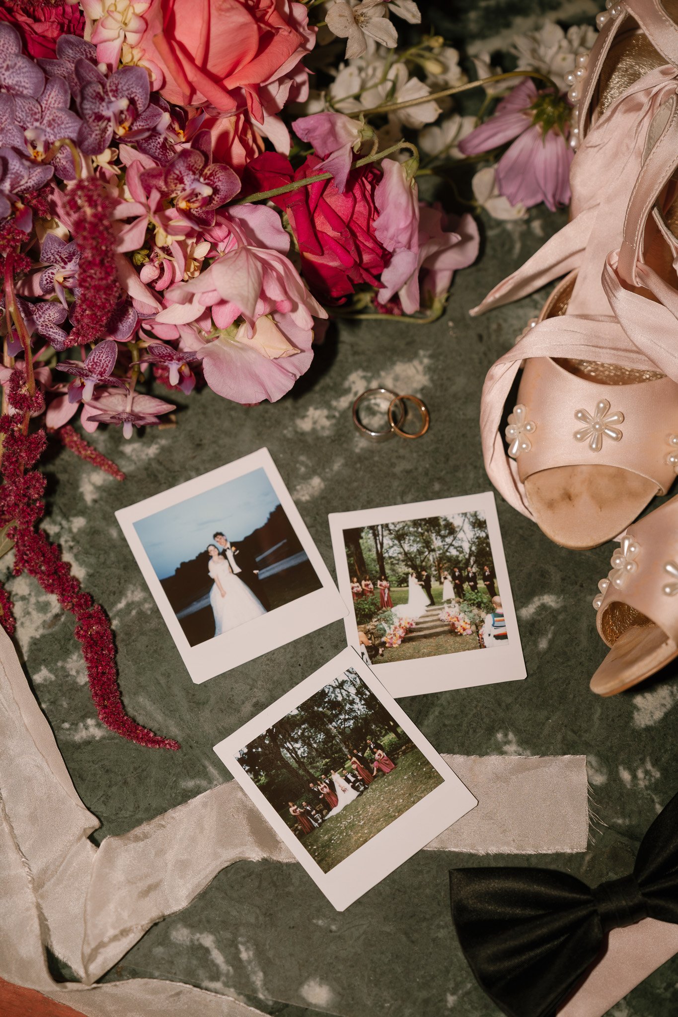 Wedding items on a table including pink flowers, wedding rings, three instant photos of a wedding couple, pink high-heeled shoes with pearl decorations, and a black bow tie.