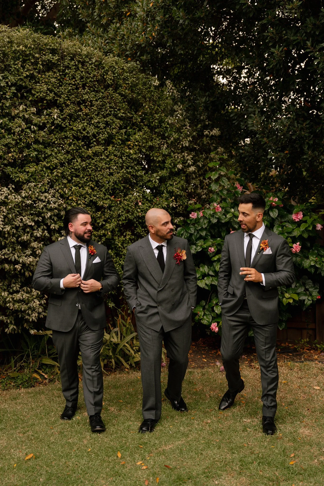 Three men in gray suits with black ties walking and talking in a garden with green bushes and pink flowers.