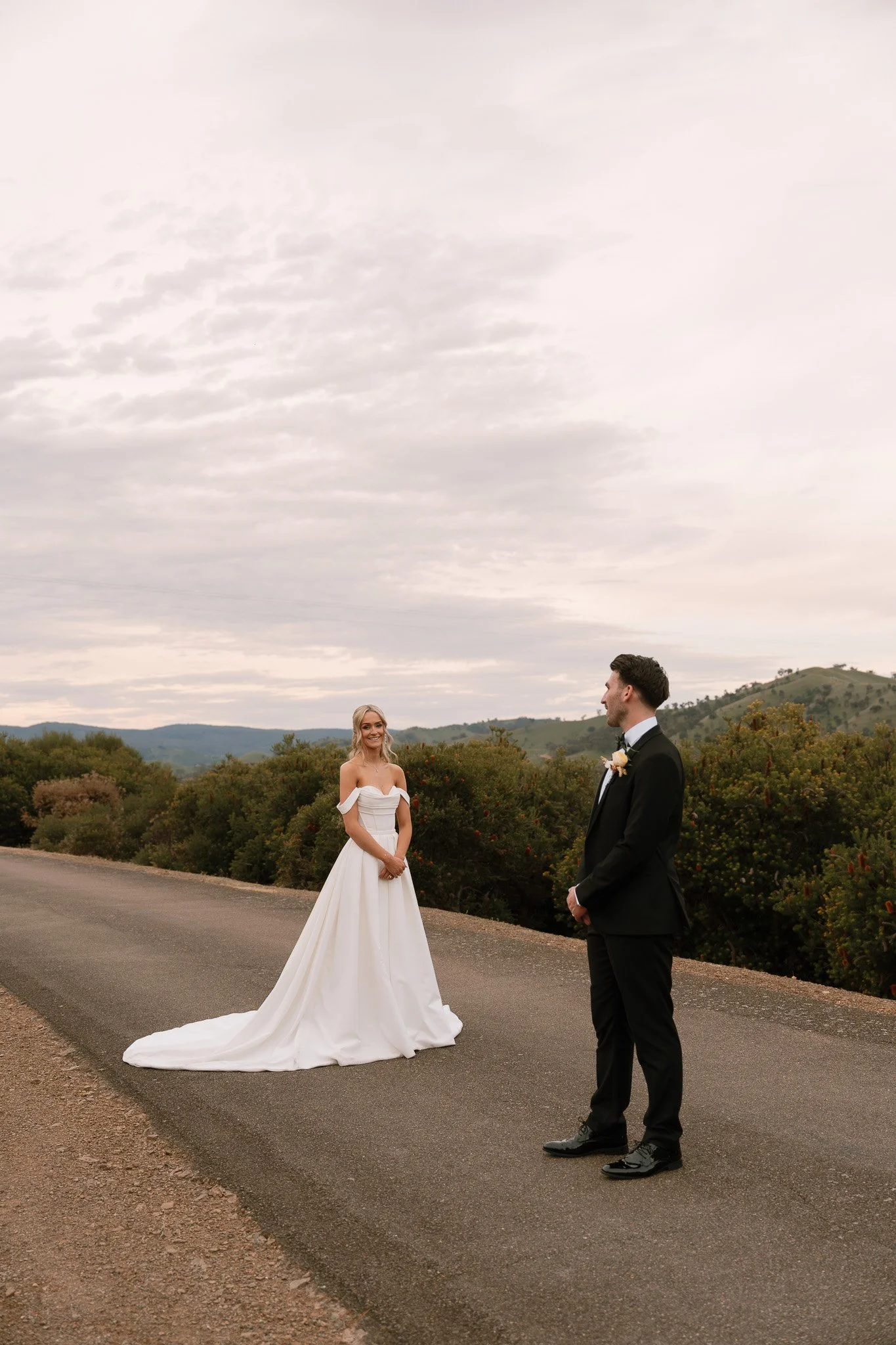 Bride in a white wedding dress and groom in a black tuxedo standing on a rural road with hills and trees in the background.