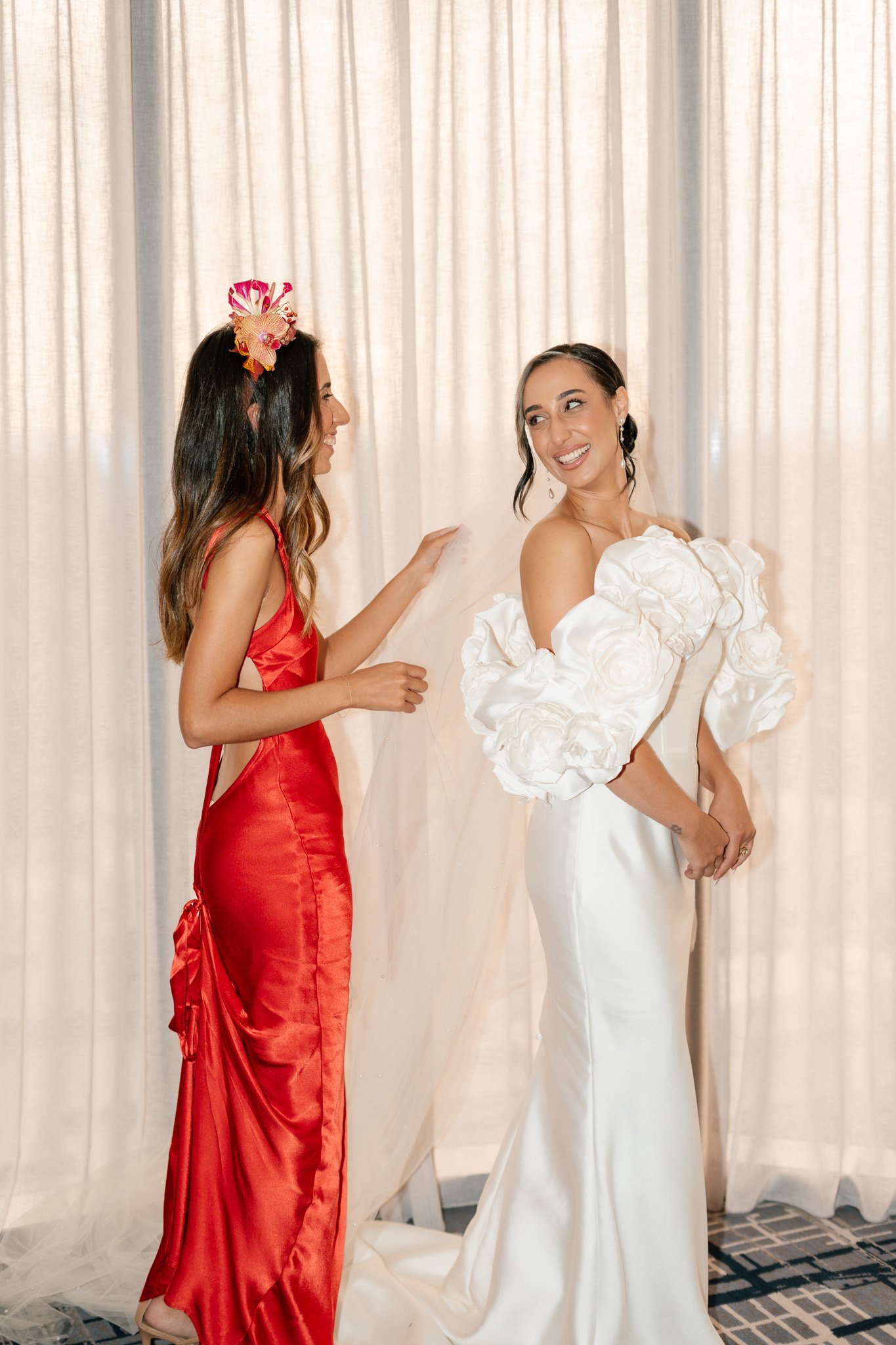 A woman in a white wedding dress with floral details on the sleeves smiling as she gets ready for her wedding, with a woman in a red dress helping her.
