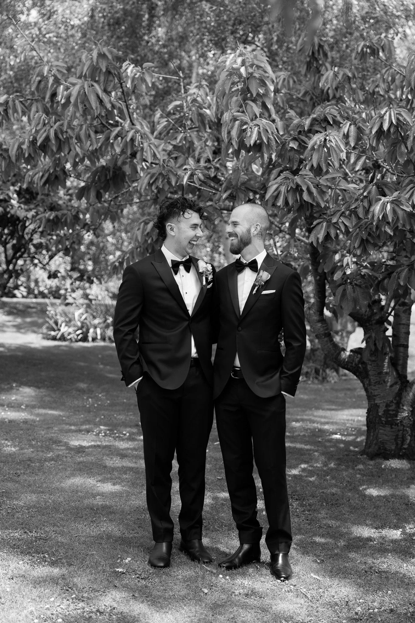 Two men in tuxedos with boutonnières standing outdoors under a large tree, smiling at each other, at a wedding or formal event.