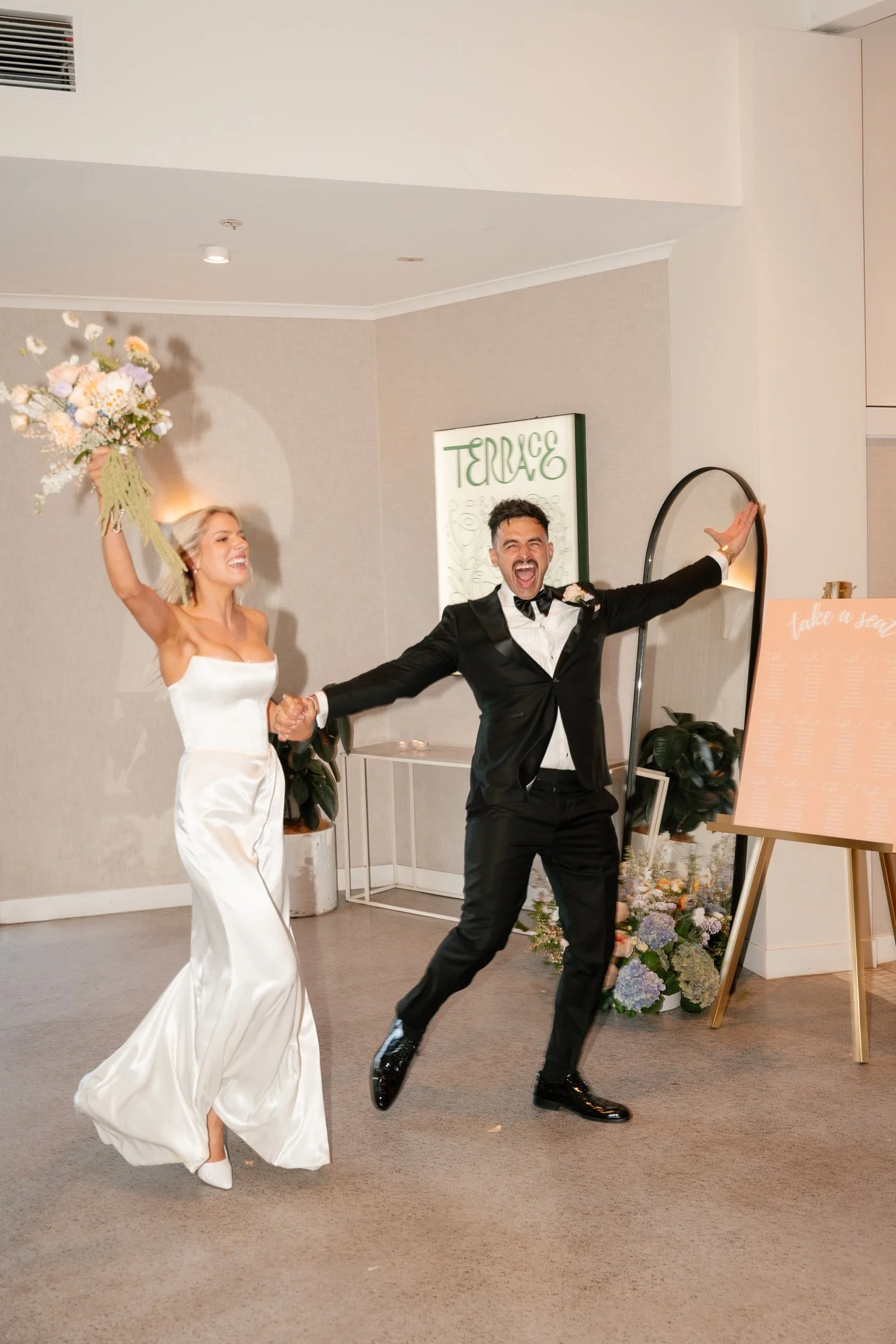 A bride and groom celebrating at their wedding reception. The bride is holding a bouquet and the groom is jumping with arms outstretched.