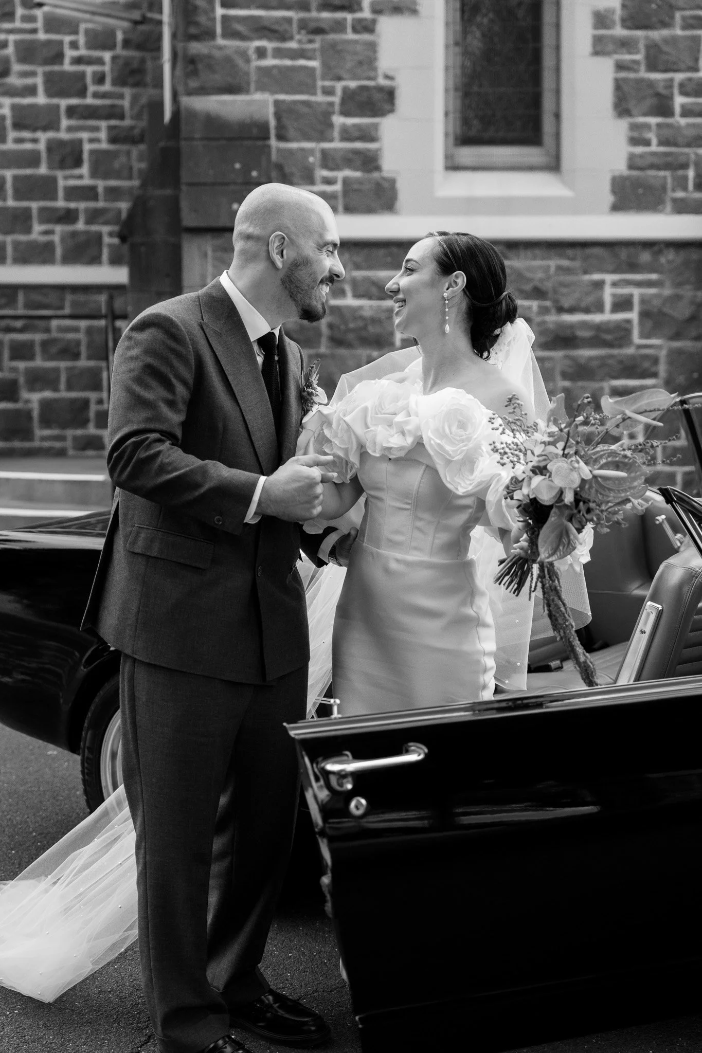 Black and white photo of a bride and groom smiling at each other, standing outside next to a vintage black car, with a brick building in the background.