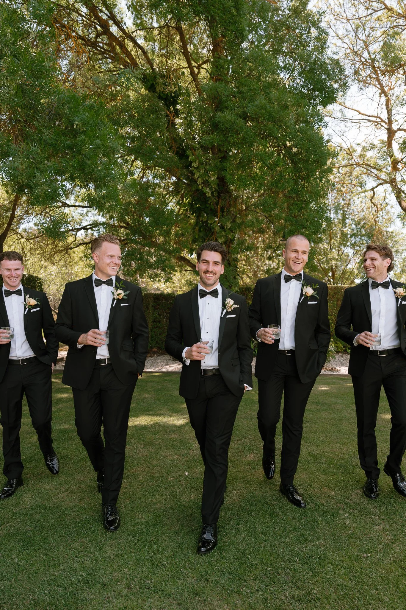 Group of men in tuxedos walking outdoors on green grass, smiling and holding glasses.
