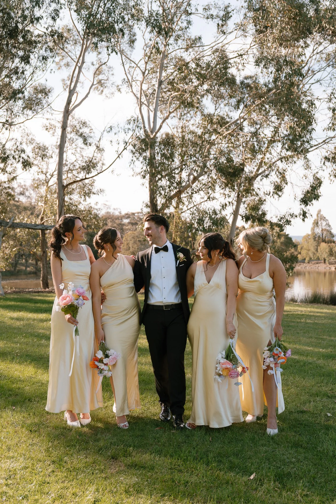 A groom in a tuxedo walking with four bridesmaids in yellow dresses holding bouquets, outdoors with trees and a body of water in the background.