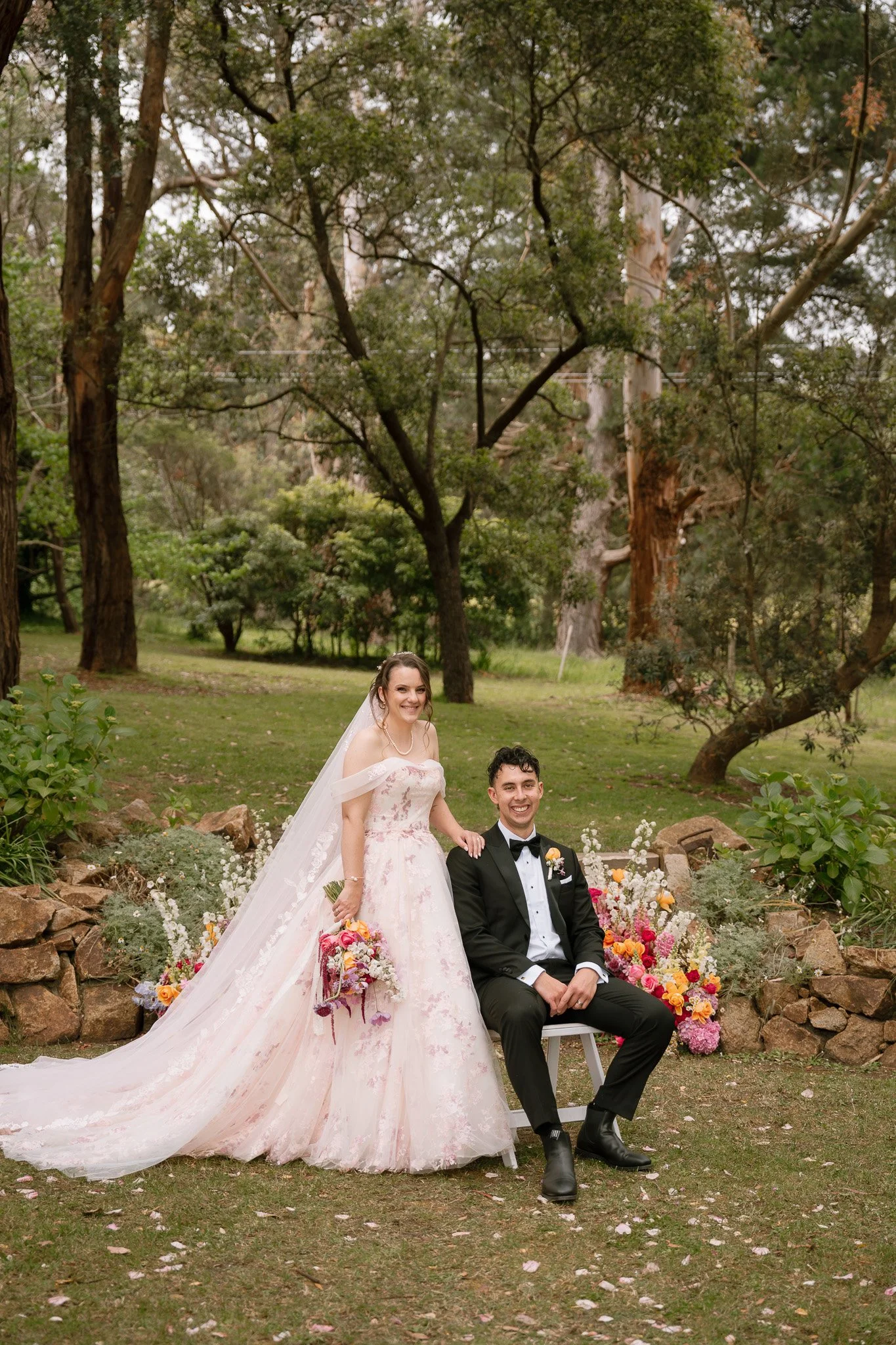 A bride and groom posing outdoors among trees and flowers on their wedding day. The bride wears a white off-shoulder gown with a long train and a veil, holding a bouquet of colorful flowers. The groom is seated, dressed in a black tuxedo with a bow t