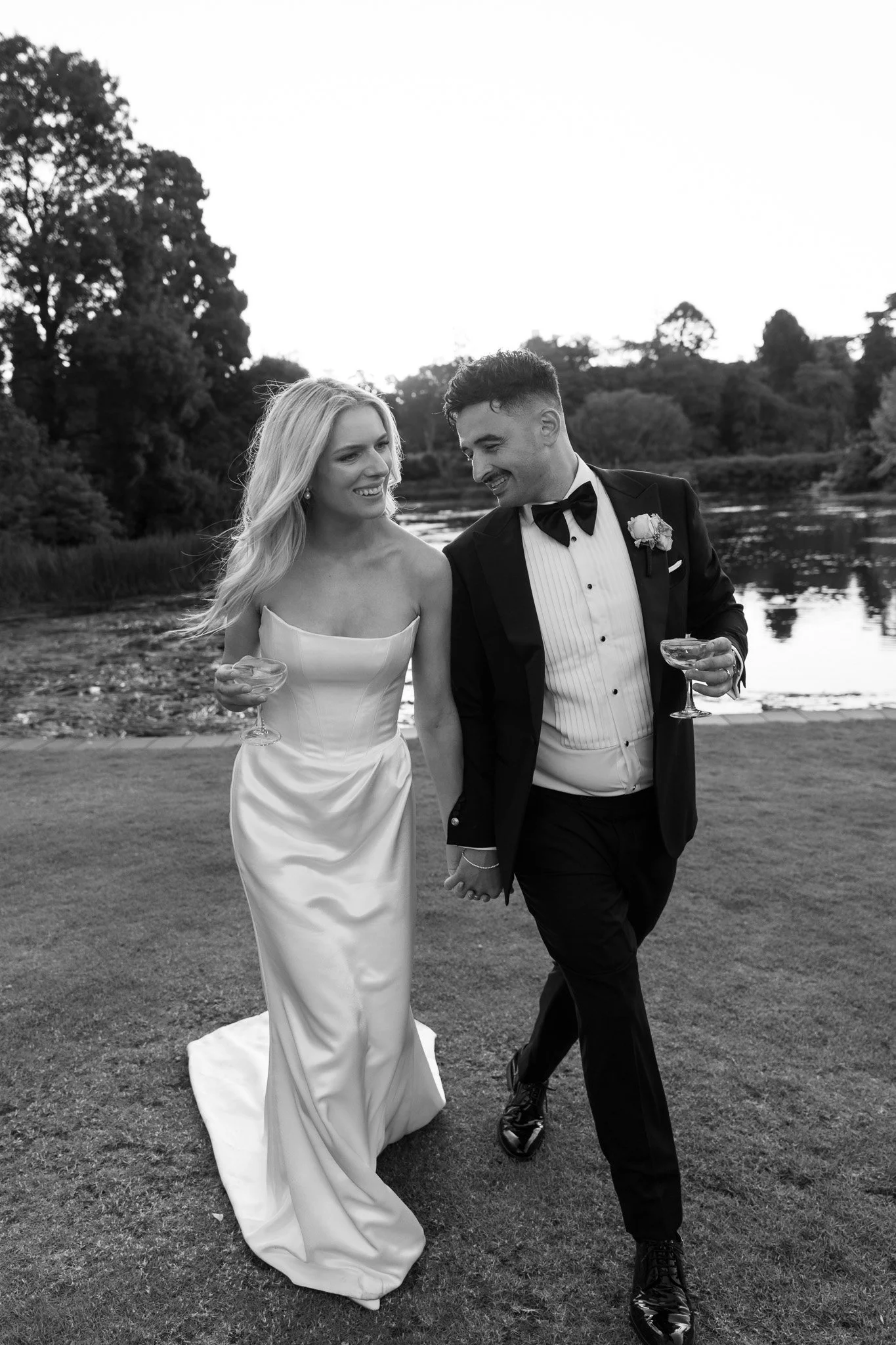 A black-and-white photo of a newlywed couple walking outdoors by a riverside, holding hands, and smiling while carrying champagne glasses.