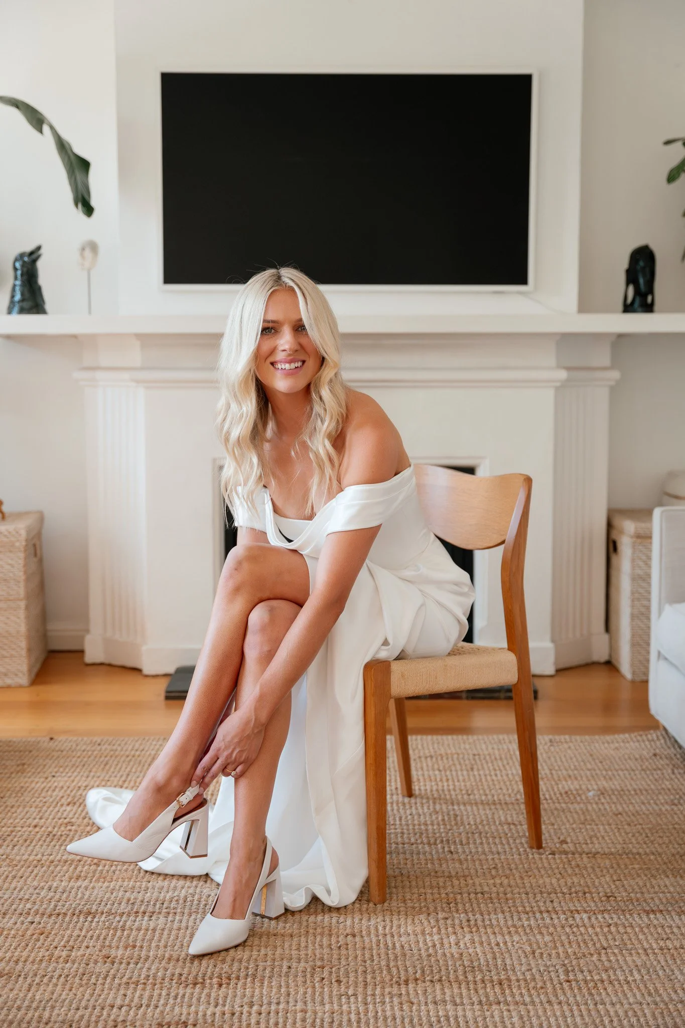 Woman in a white dress sitting on a wooden chair, adjusting her white high heels, smiling in a modern living room with a black TV mounted on a white fireplace mantel in the background.