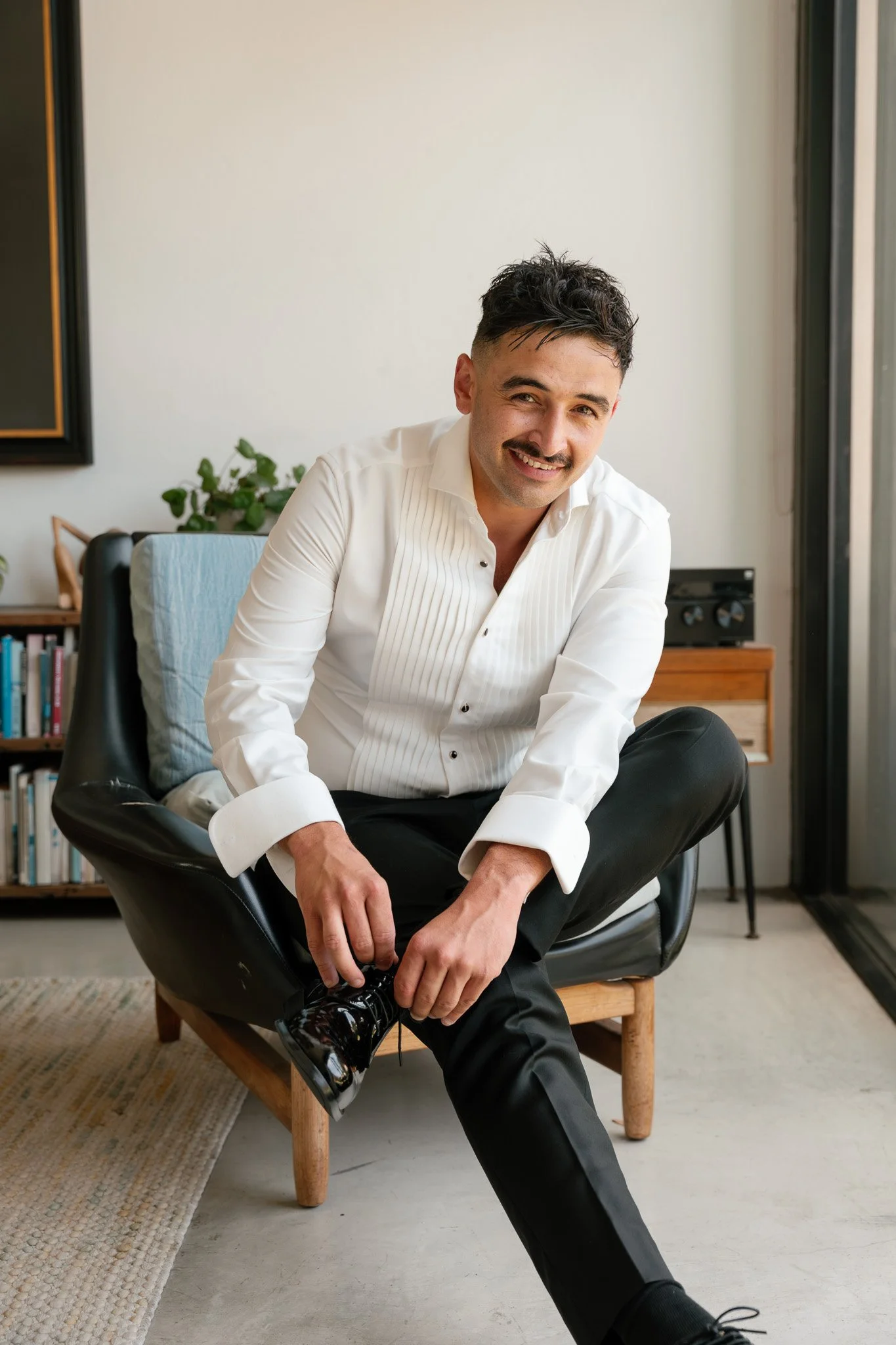 A man with dark hair and a mustache wearing a white shirt and black pants, sitting in a modern living room, smiling and tying his black shoes.