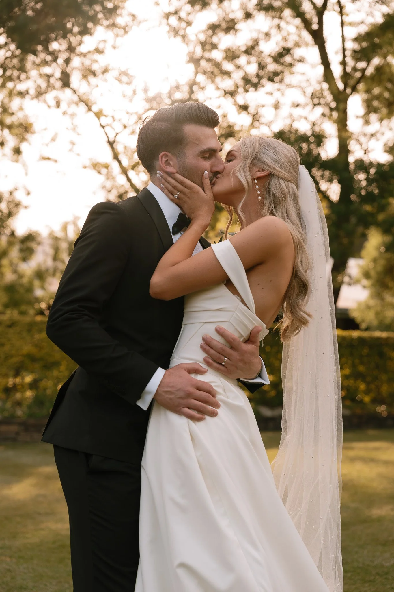 A bride and groom kiss outdoors at sunset, with trees in the background.
