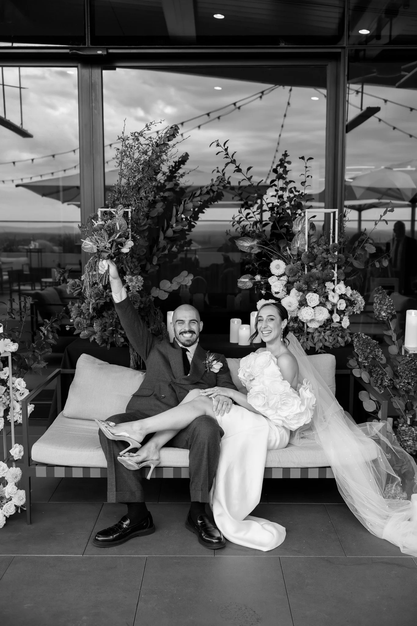 Black and white photo of a newlywed couple sitting on a couch after their wedding ceremony, surrounded by floral arrangements and candles, celebrating outdoors.