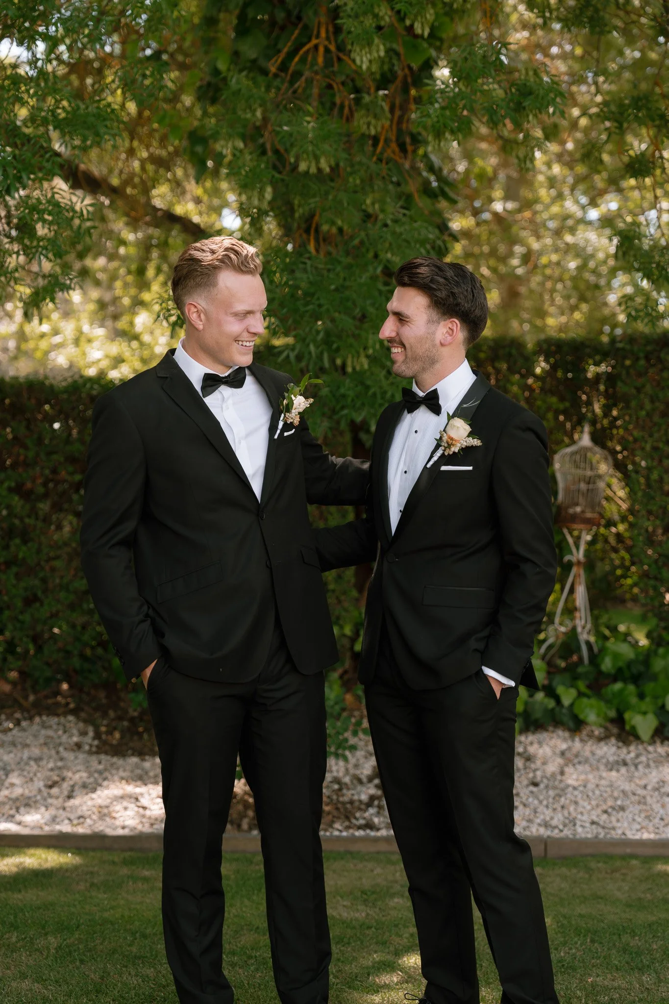 Two men in black tuxedos with bow ties and boutonnières, standing outdoors in front of a green leafy background, smiling at each other.