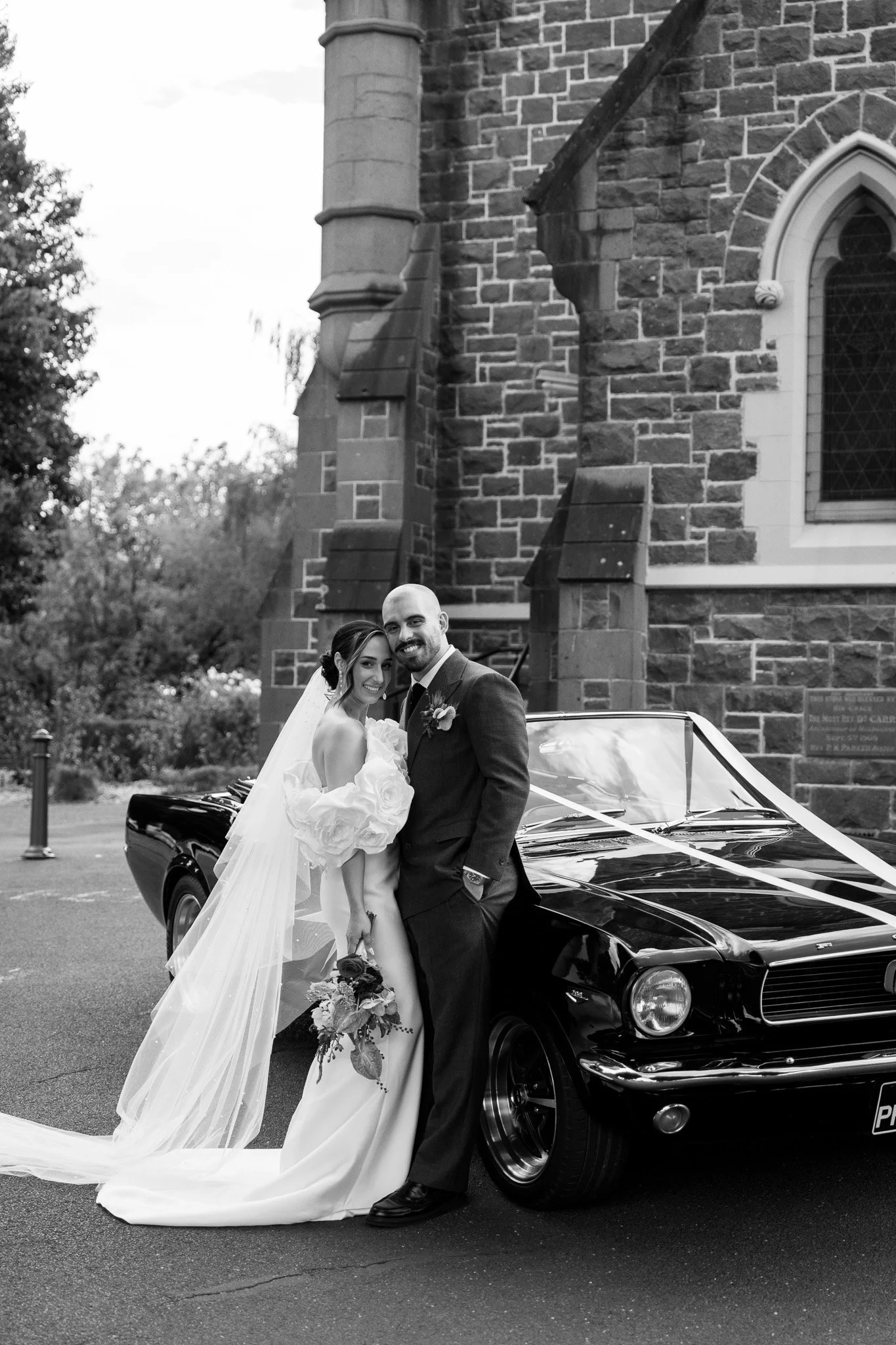 A bride and groom posing beside a vintage black car decorated with a white ribbon in front of a stone church.