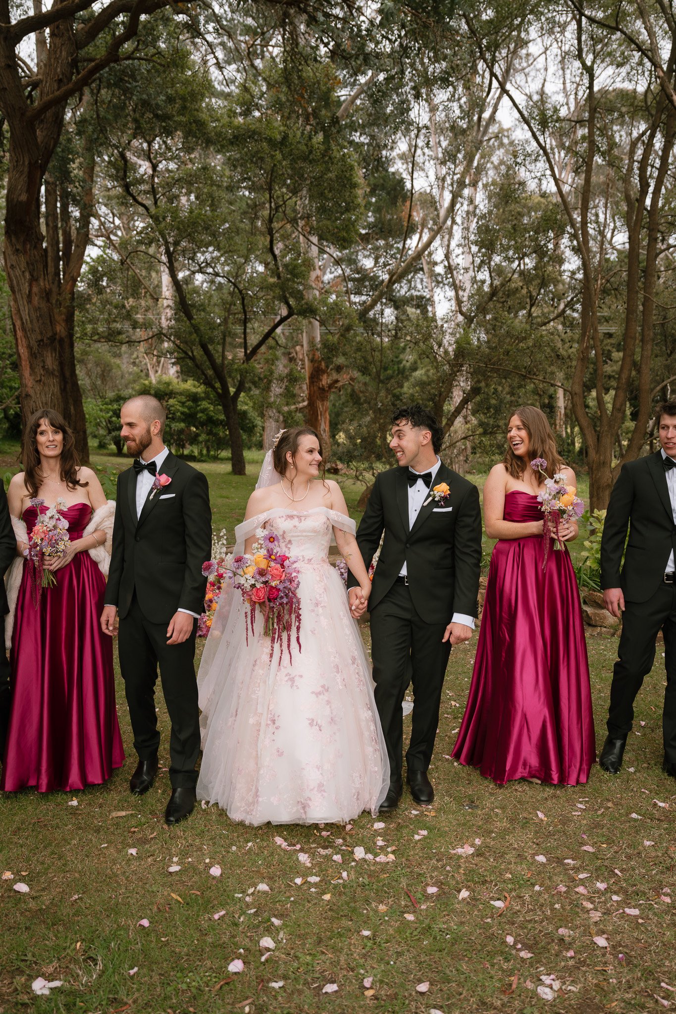 A wedding party walking outdoors in a park with trees, including a bride in a white gown, a groom in a black tuxedo, and bridesmaids in matching magenta dresses, holding bouquets.