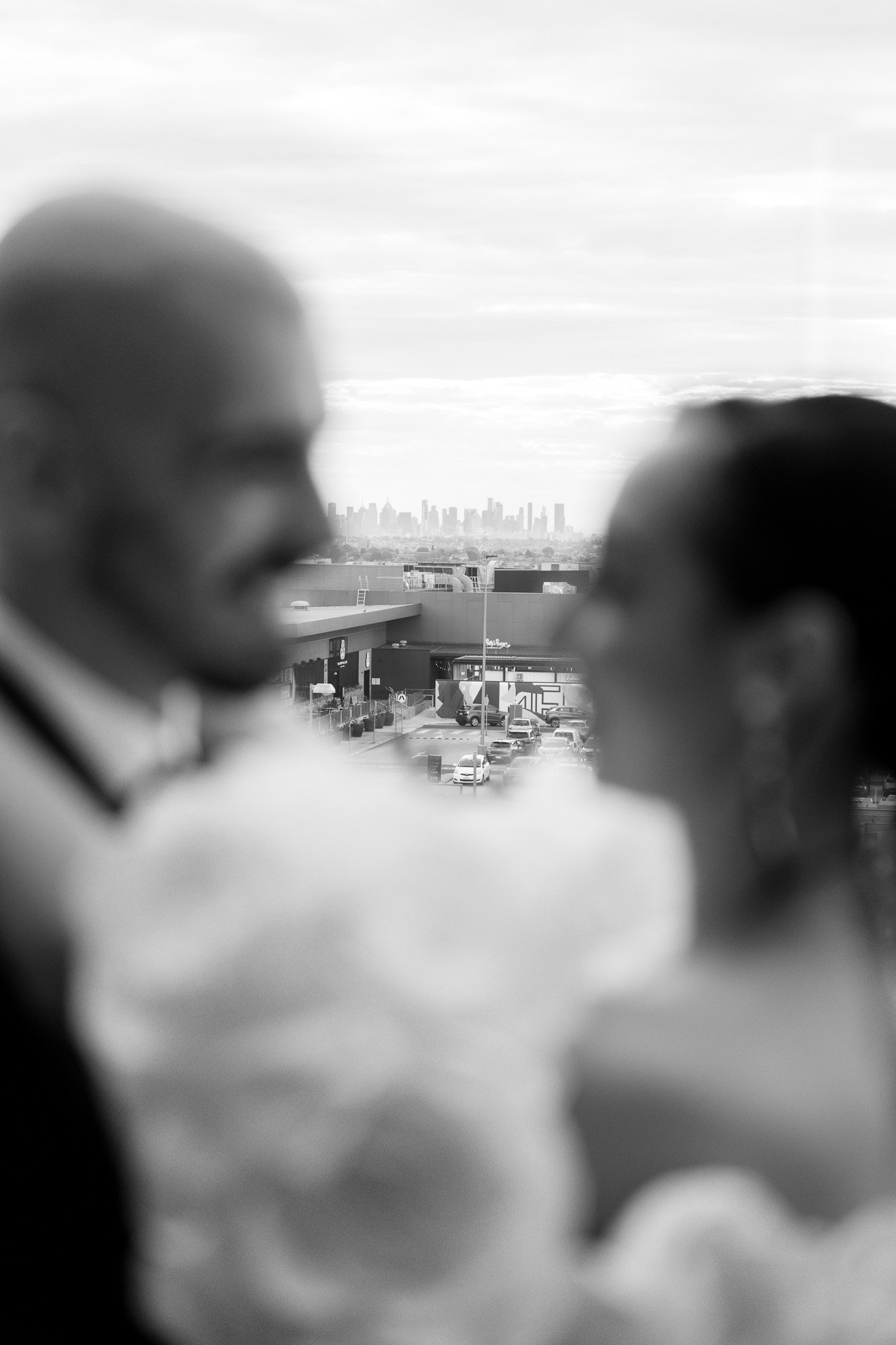 Blurry black and white photo of a man and woman in focus with a cityscape and parking lot visible through a window in the background.