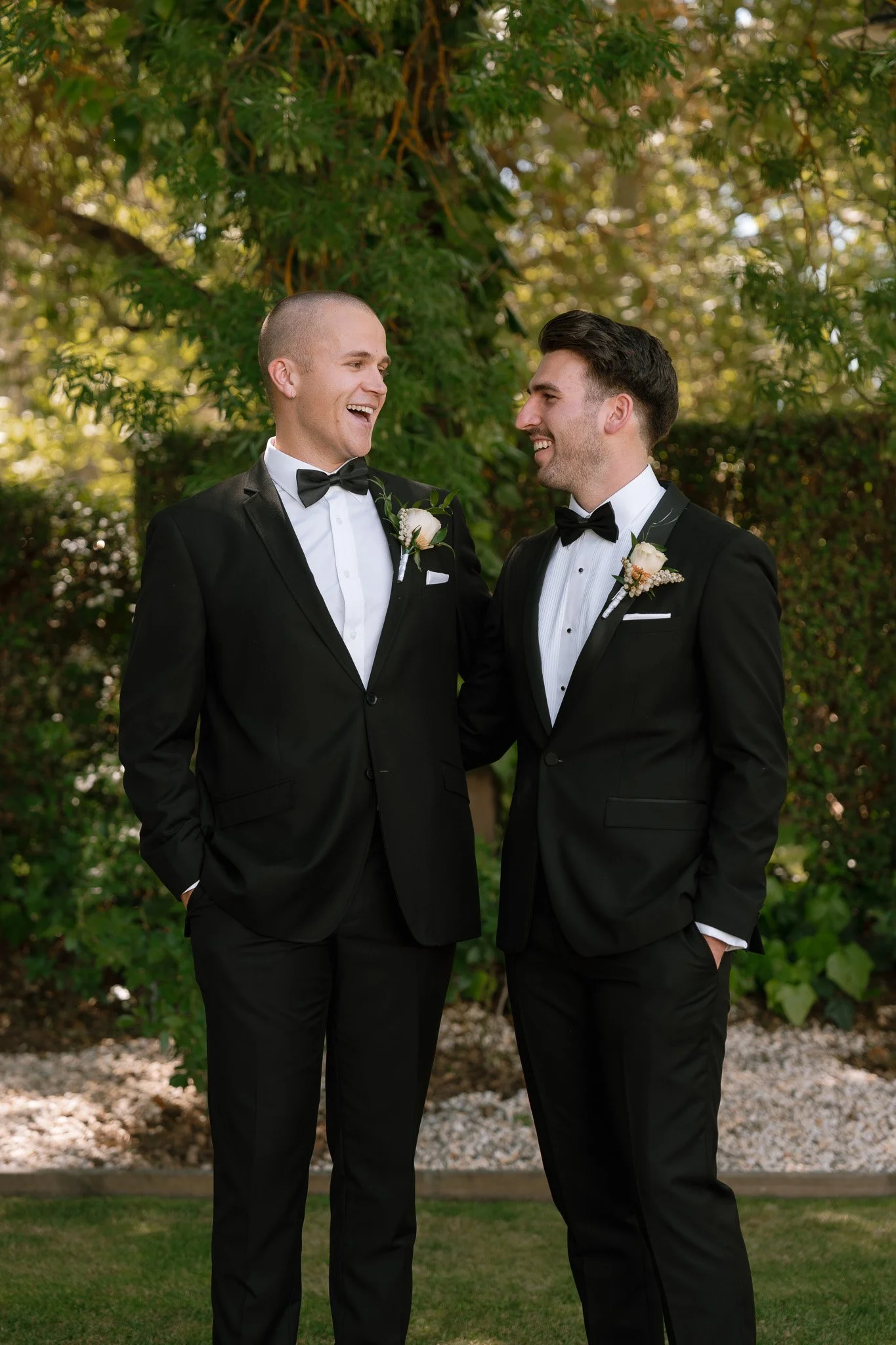 Two men in tuxedos with boutonnieres, smiling and sharing a moment outdoors.