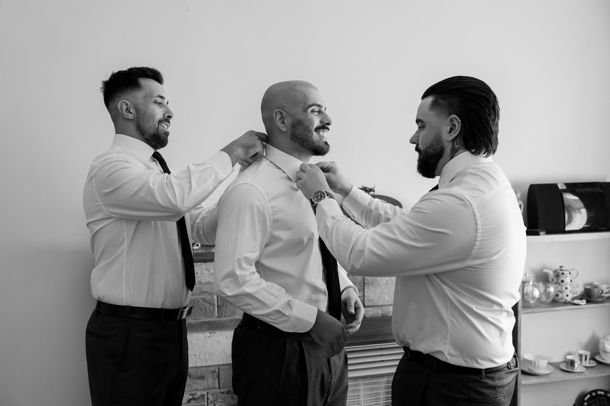 Three men dressed in white shirts and black ties preparing for a formal event, with two men adjusting the third man's collar and tie.