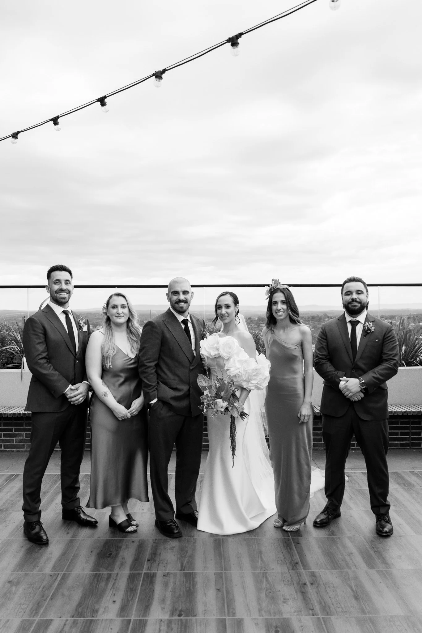 Black and white photo of a wedding party with three men and three women standing on a rooftop terrace, with a scenic view of the landscape in the background. The bride is holding a bouquet of flowers, and everyone is dressed in formal attire.